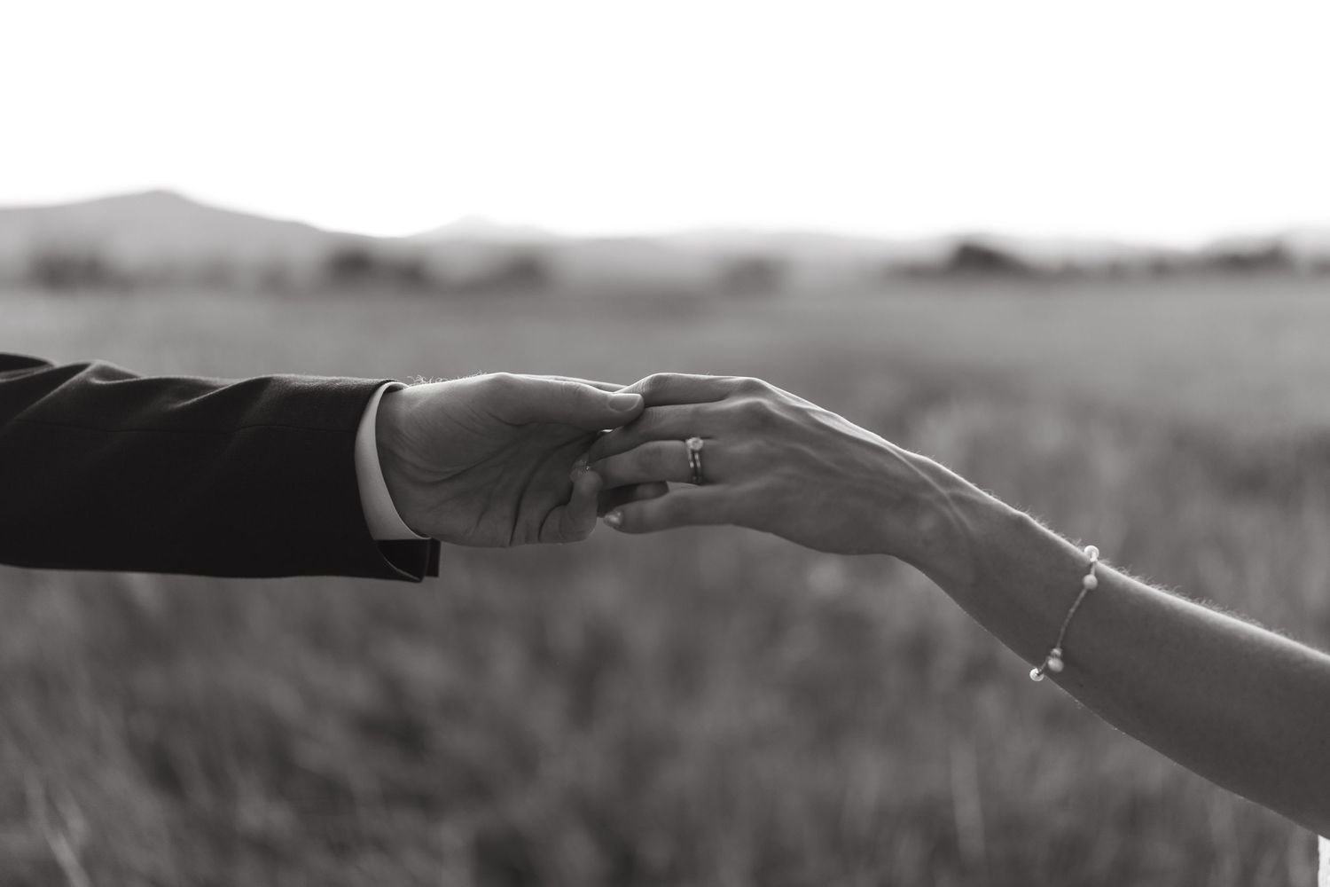 A black and white close-up of two hands reaching for each other in an outdoor setting.