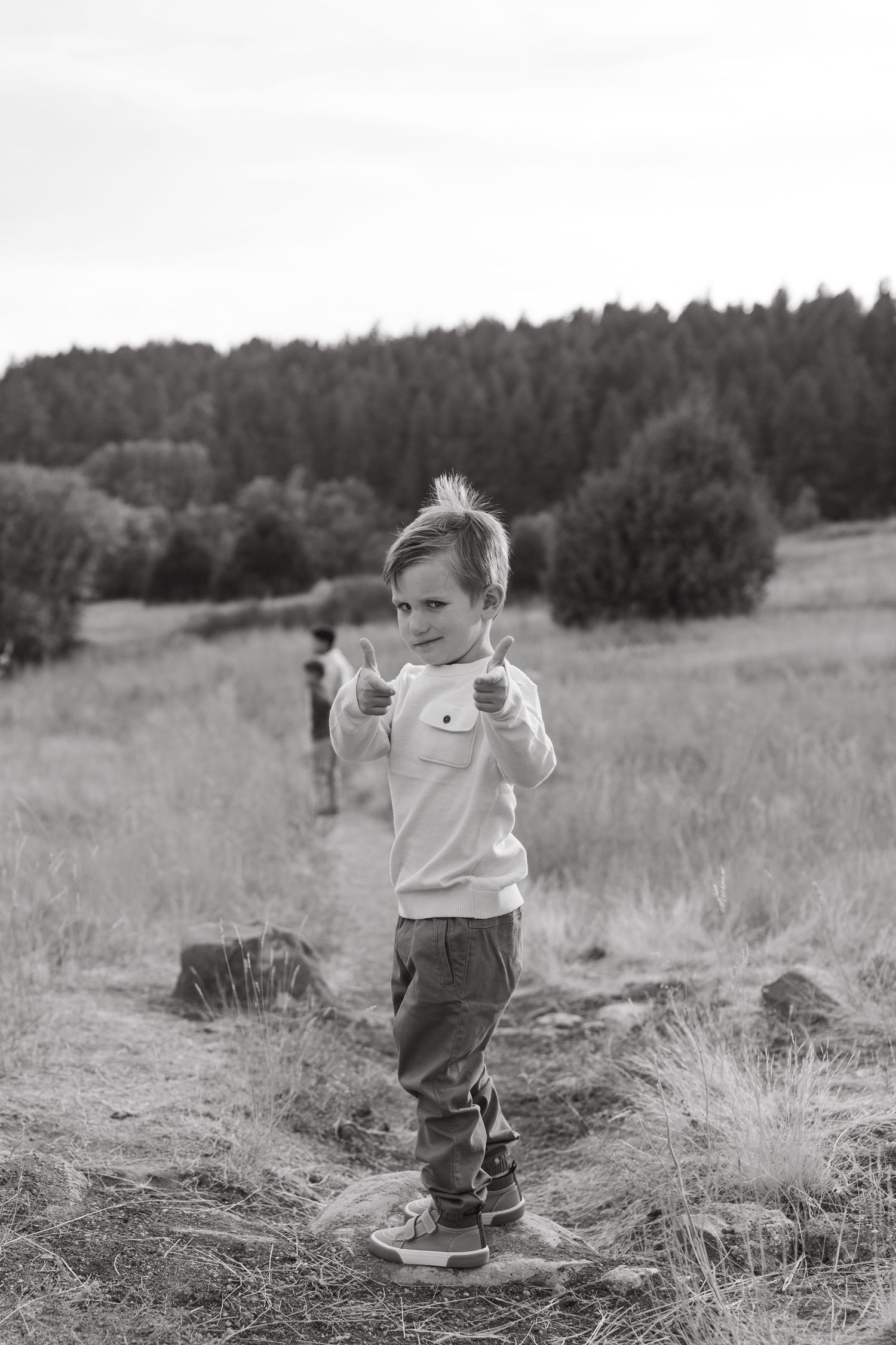 A black and white photograph of a child posing confidently in a rural field with mountains in the background.