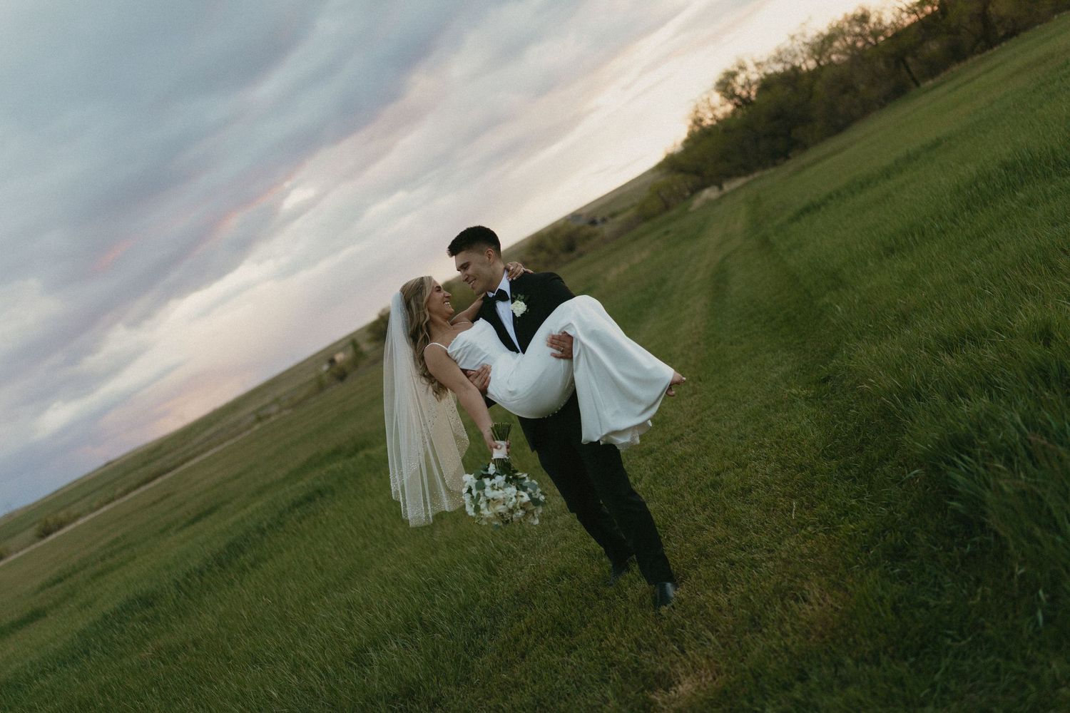 Groom carries bride in wedding attire across lush green field at sunset with dramatic pink sky backdrop.