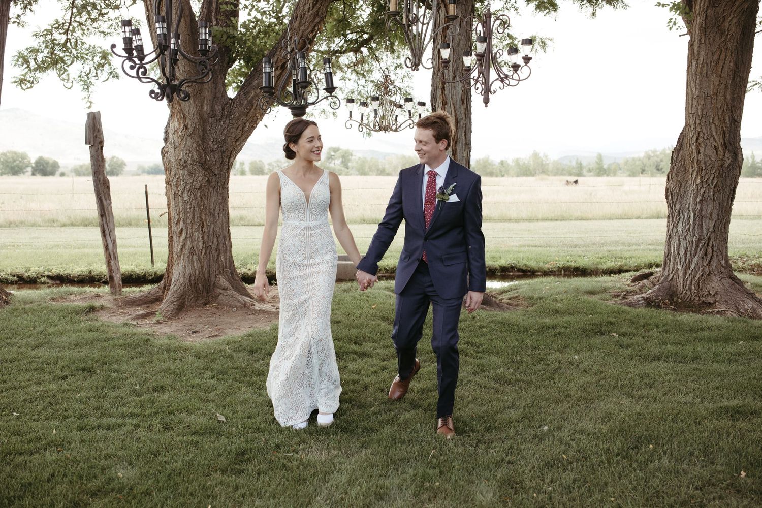 Wedding couple walks hand in hand under tall trees in a grassy outdoor setting.