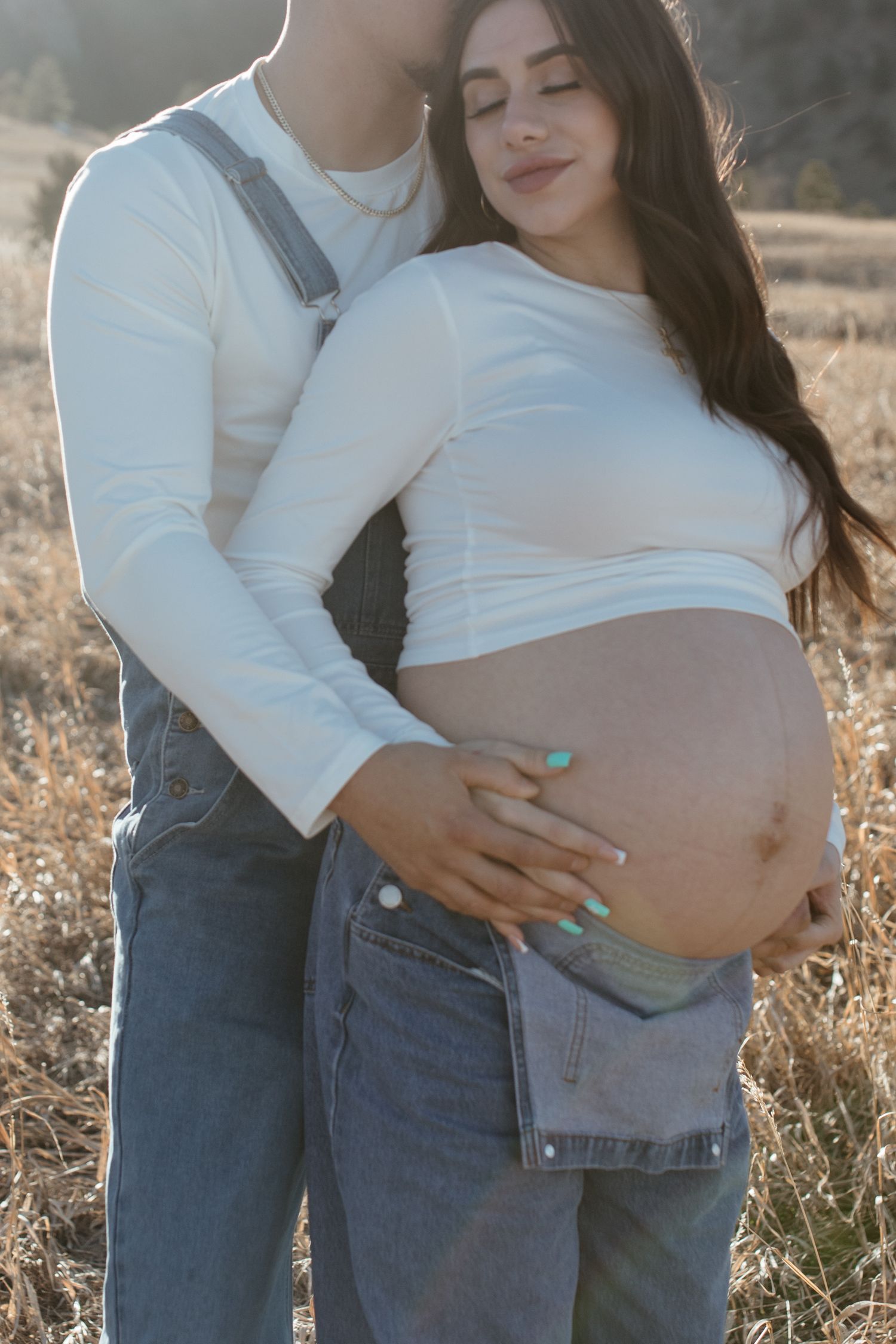 Romantic maternity photo showing couple in white tops and denim embracing in sunlit field.