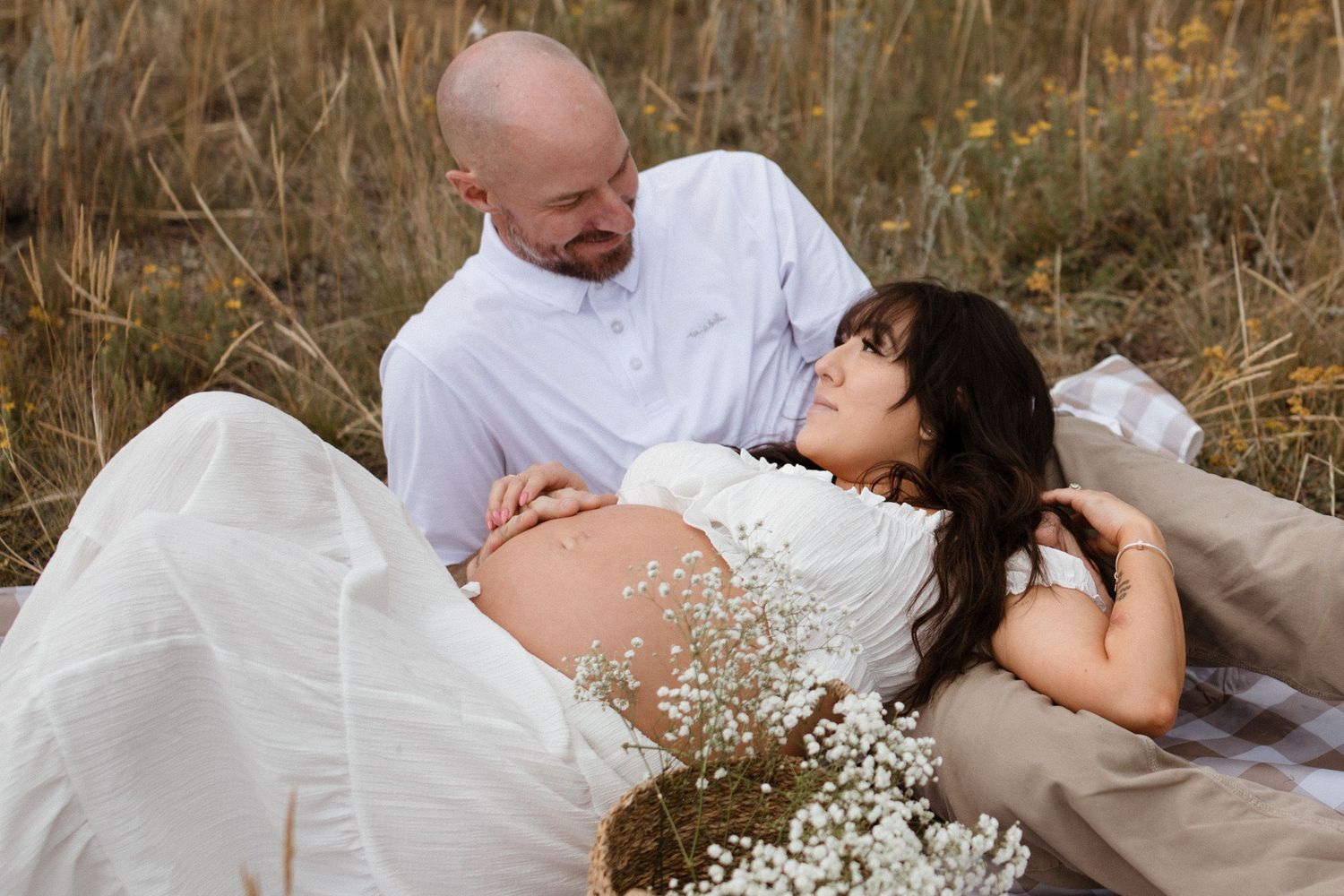 Couple in white clothing lying in tall grass with baby's breath flowers during outdoor maternity photoshoot.