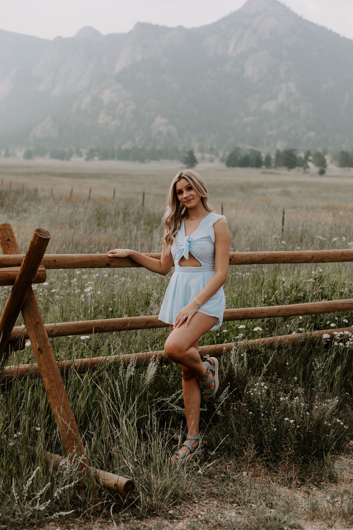 Summer fashion portrait by wooden fence with mountain landscape background wearing light blue dress.