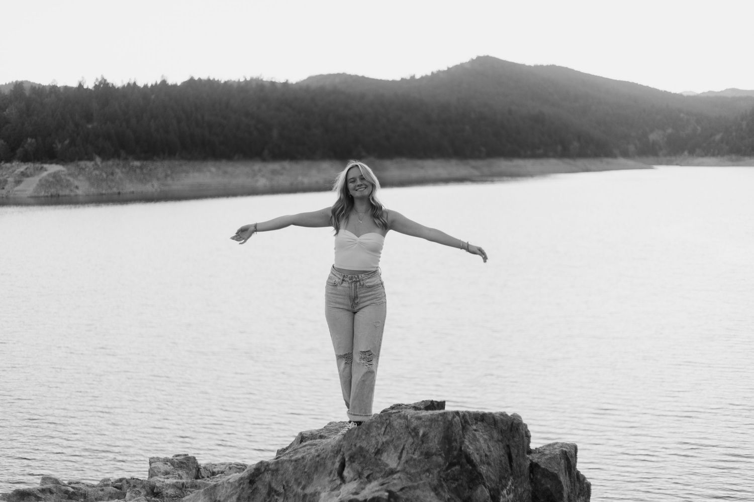 Black and white photo of person standing with arms outstretched on rocky outcrop by serene lake with mountains in background.