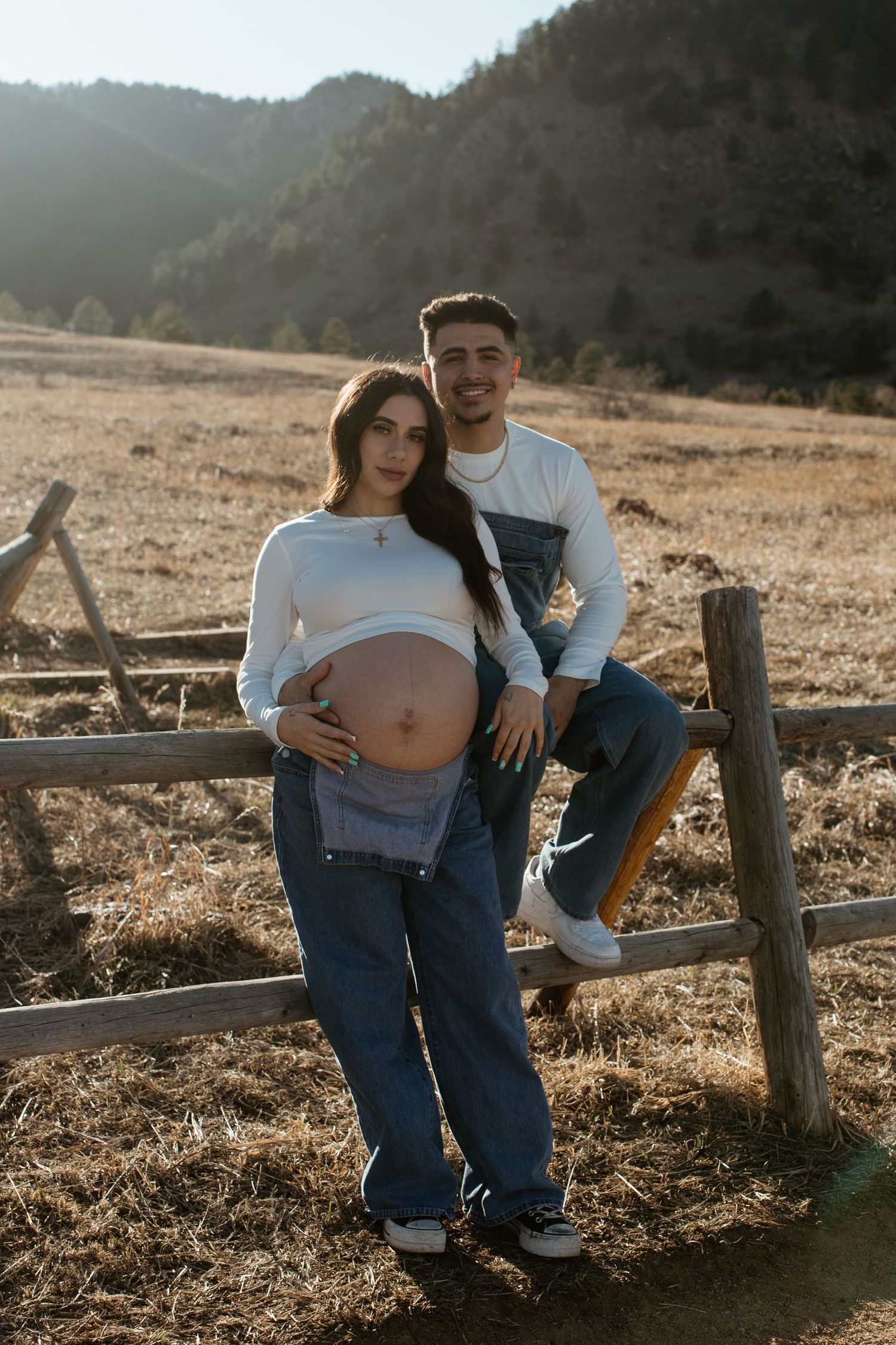 Expectant couple posing by rustic wooden fence in mountain field during sunset.