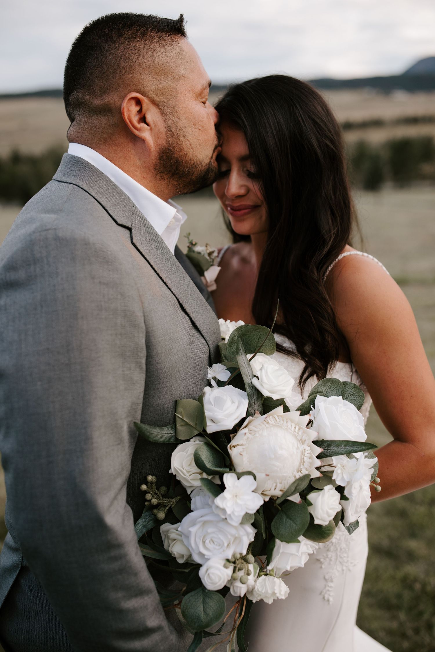 Intimate moment between newlyweds with white rose bouquet against mountain backdrop.