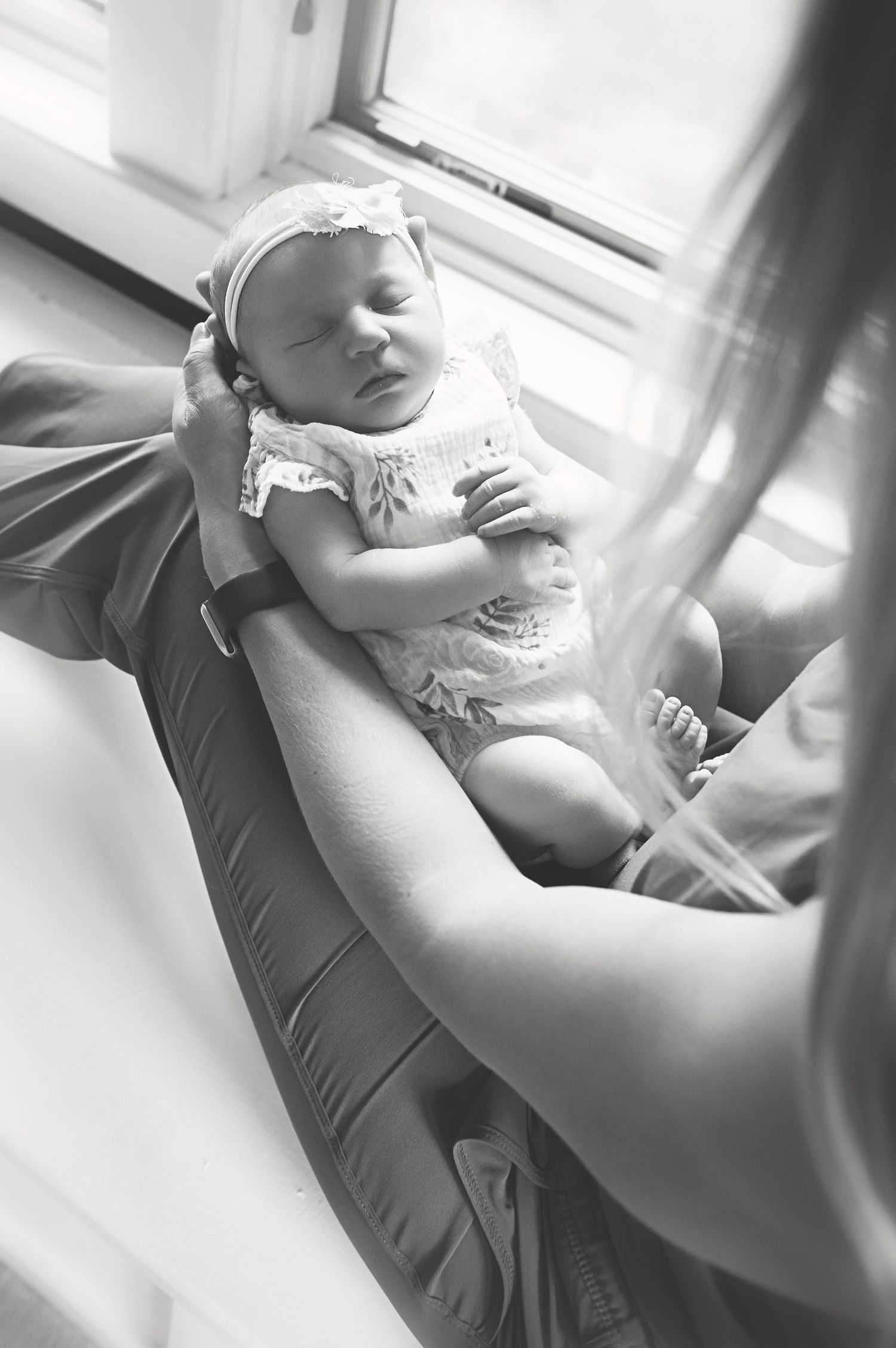 Black and white photo of sleeping newborn baby in white outfit being cradled.