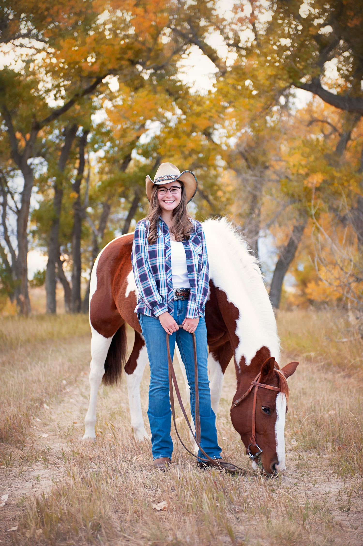 Cowgirl with plaid shirt stands next to grazing brown and white horse in fall setting with golden trees.
