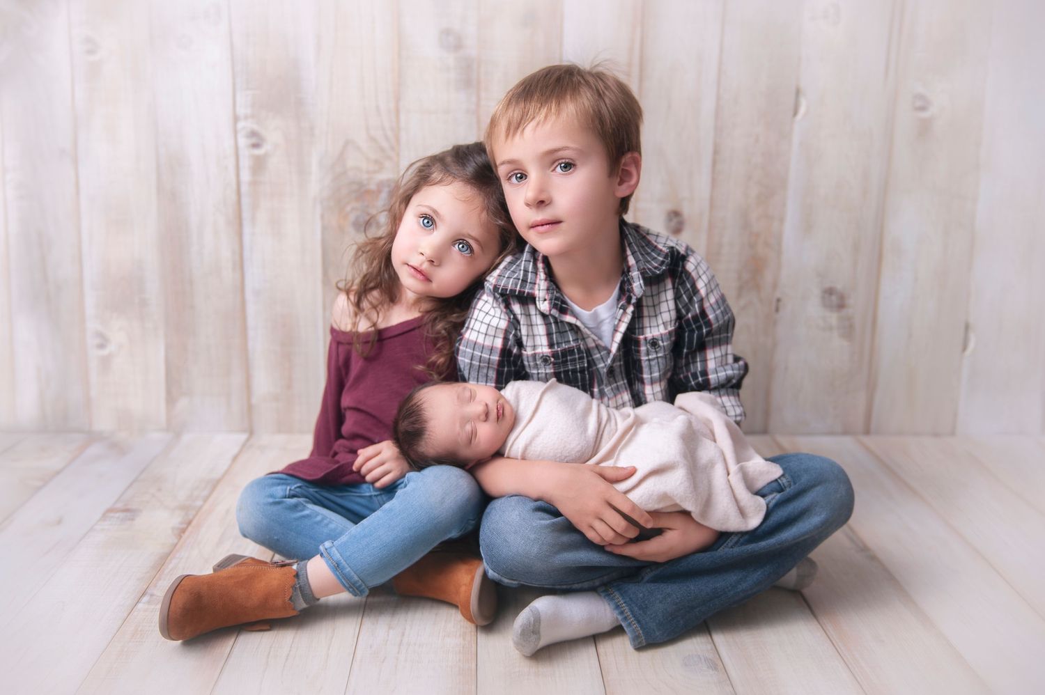 Three siblings sit together on a wooden floor with the eldest holding a newborn baby.