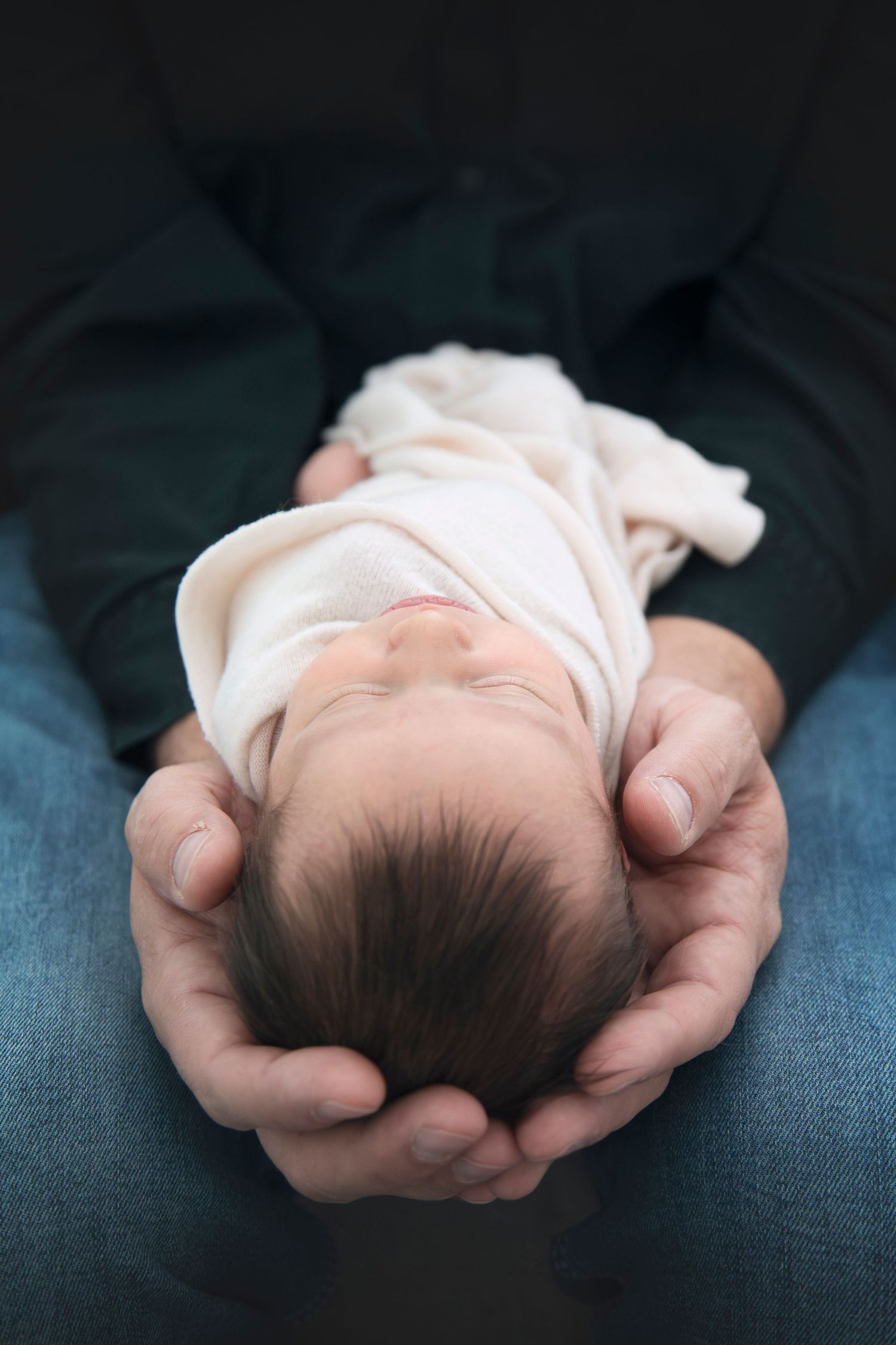 Overhead view of tiny sleeping newborn cradled in adult hands on blue surface.