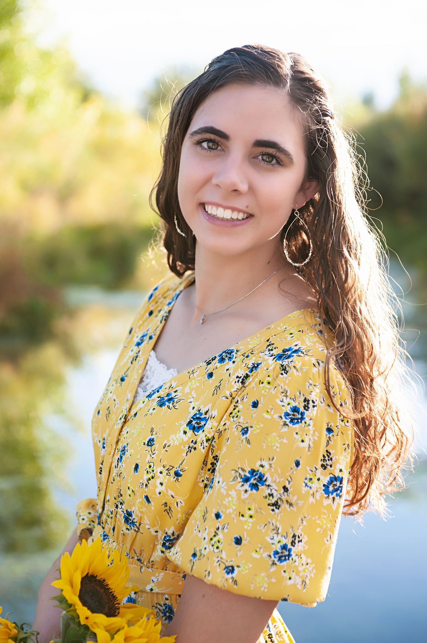 Person in yellow floral dress holding sunflower against blurred nature background.