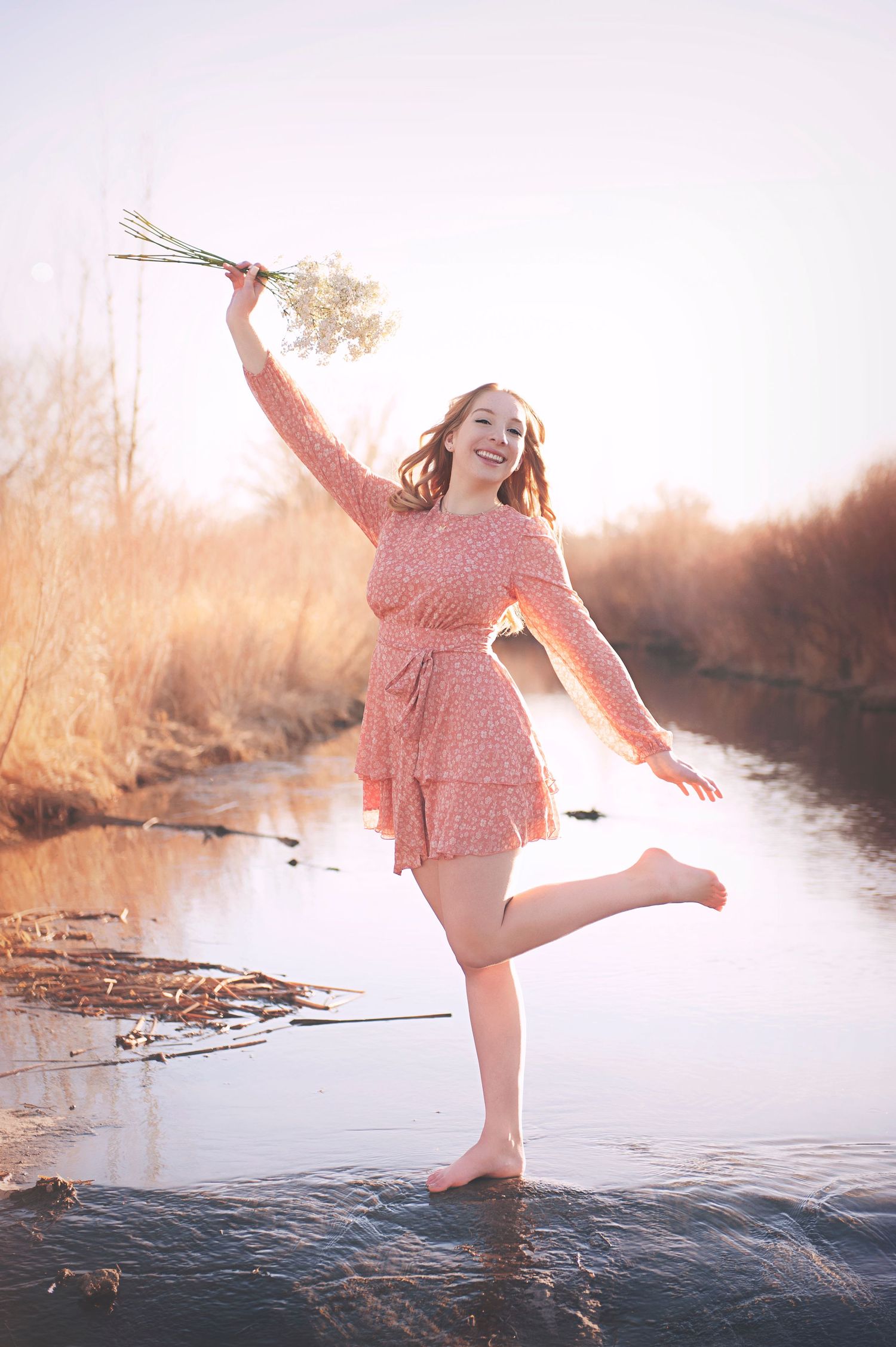 Person in pink dress splashing in water while holding flowers at sunset.