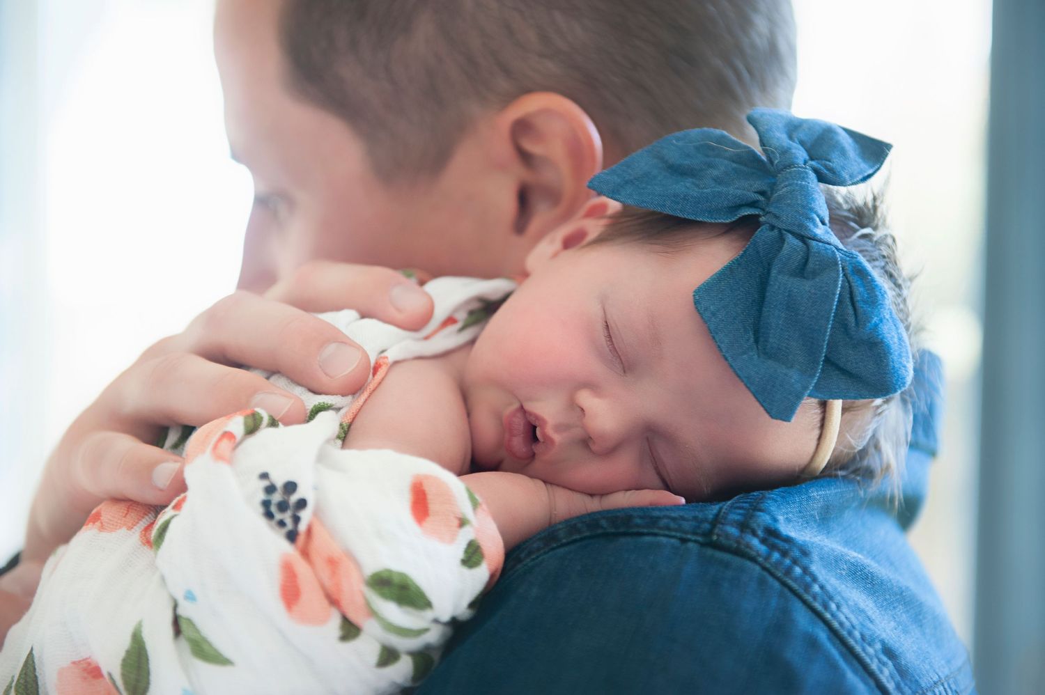 Sleeping baby in floral wrap and blue bow rests peacefully on shoulder.