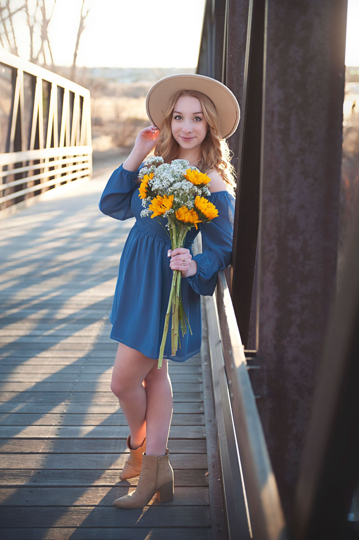 Stylish outfit with blue dress and hat while holding yellow flowers on a wooden bridge.