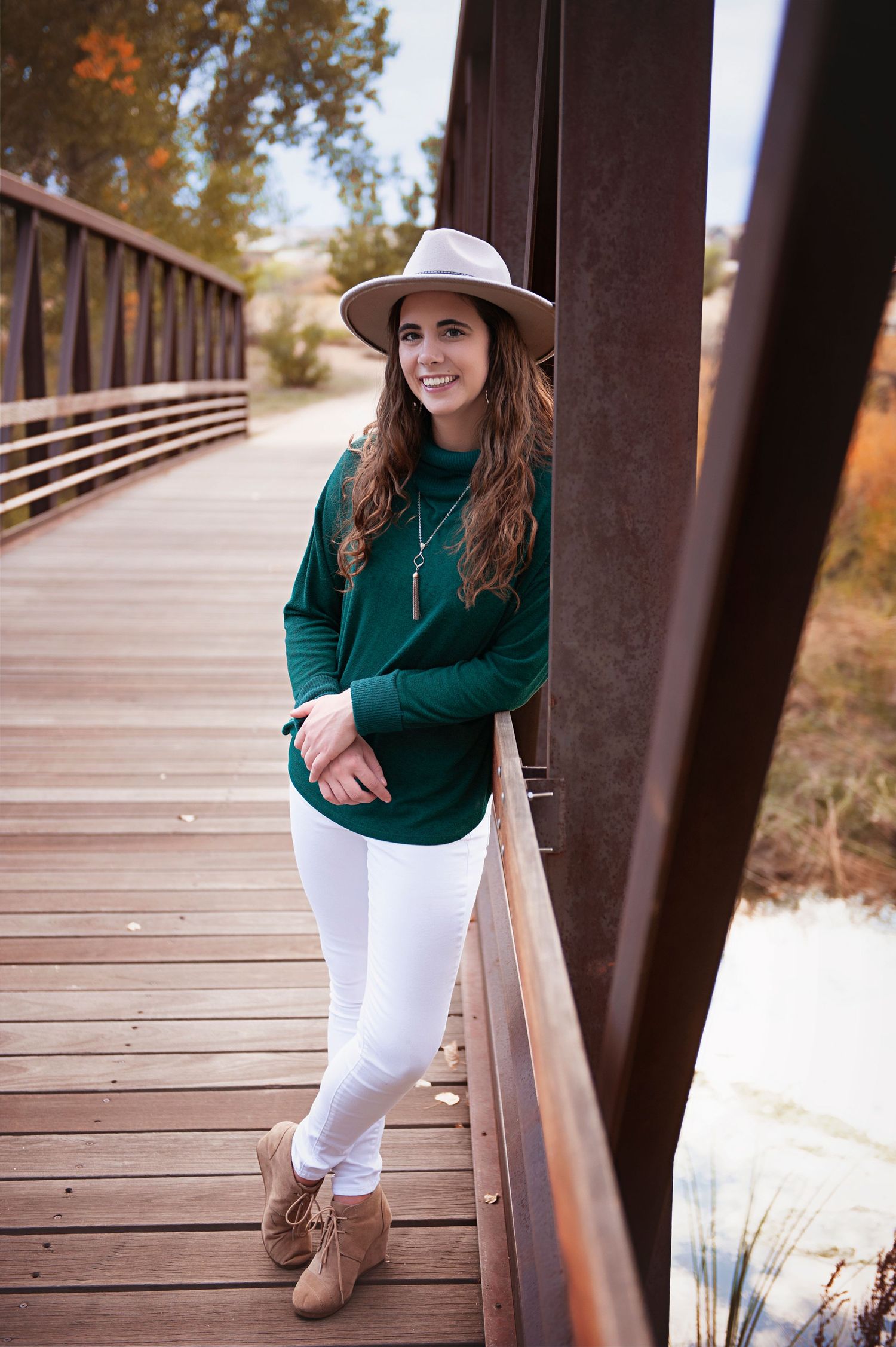 Person in green sweater and white pants posing casually against wooden bridge railing.