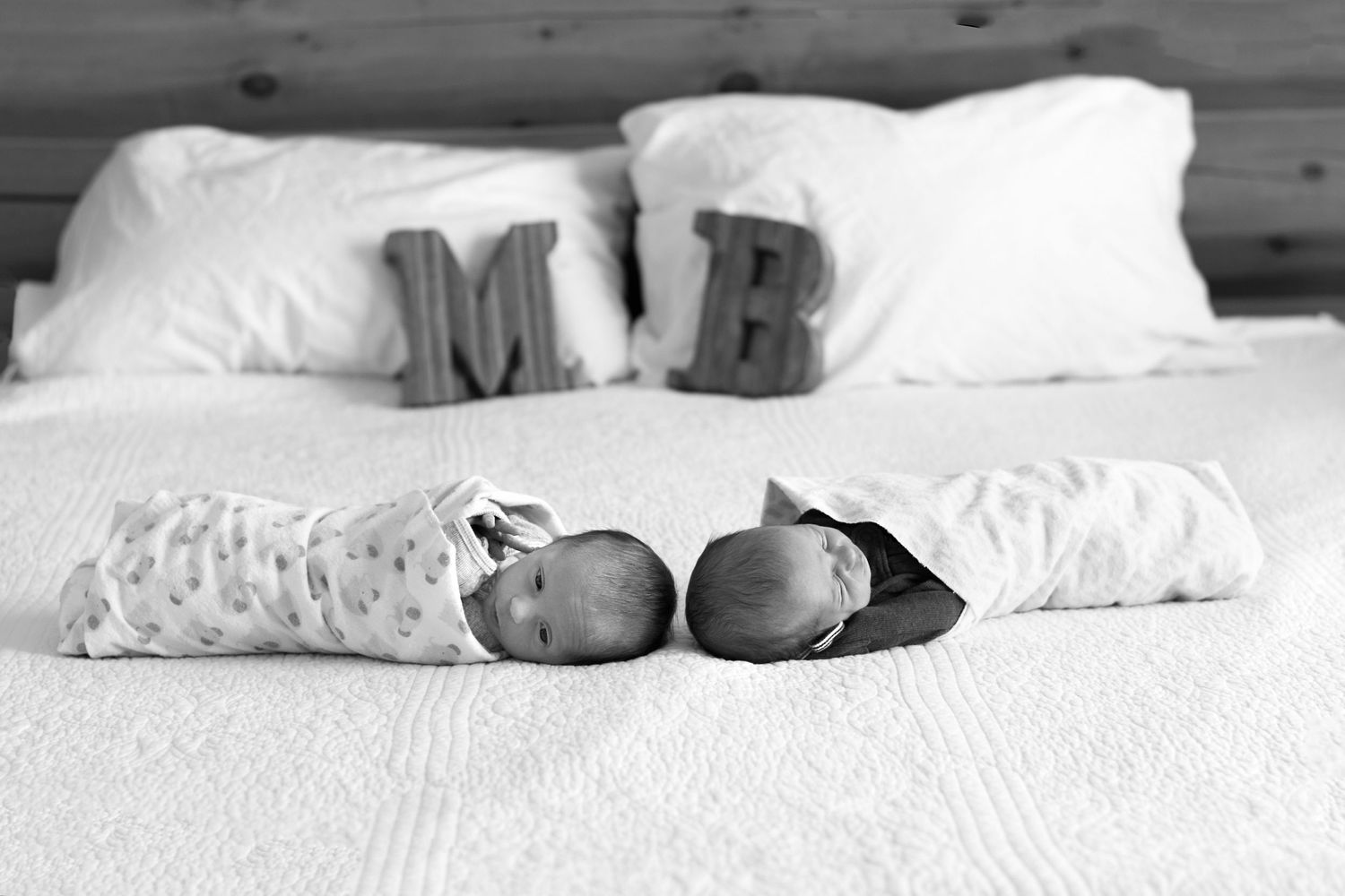 Black and white photo of twin babies sleeping on bed with decorative letters M and B on pillows.