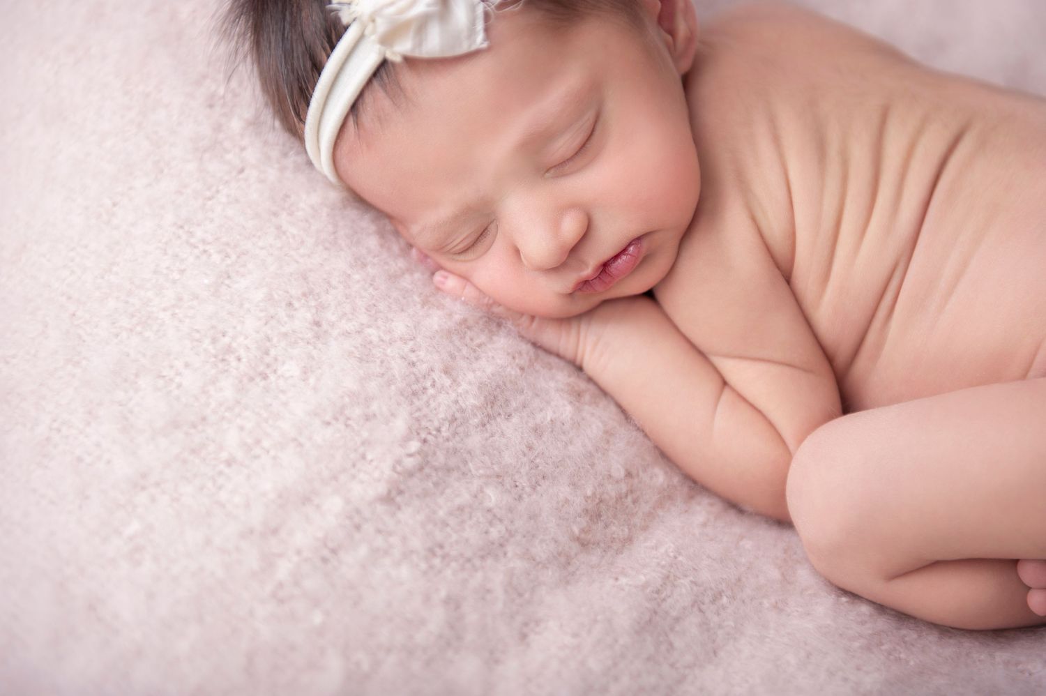 Peaceful sleeping newborn in white headband resting on soft pink textured blanket.