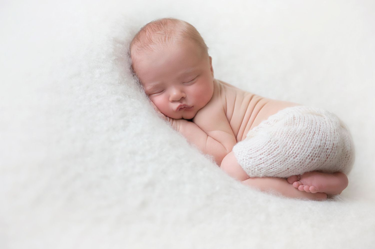 Sleeping newborn partially wrapped in white knit fabric on fluffy white backdrop.