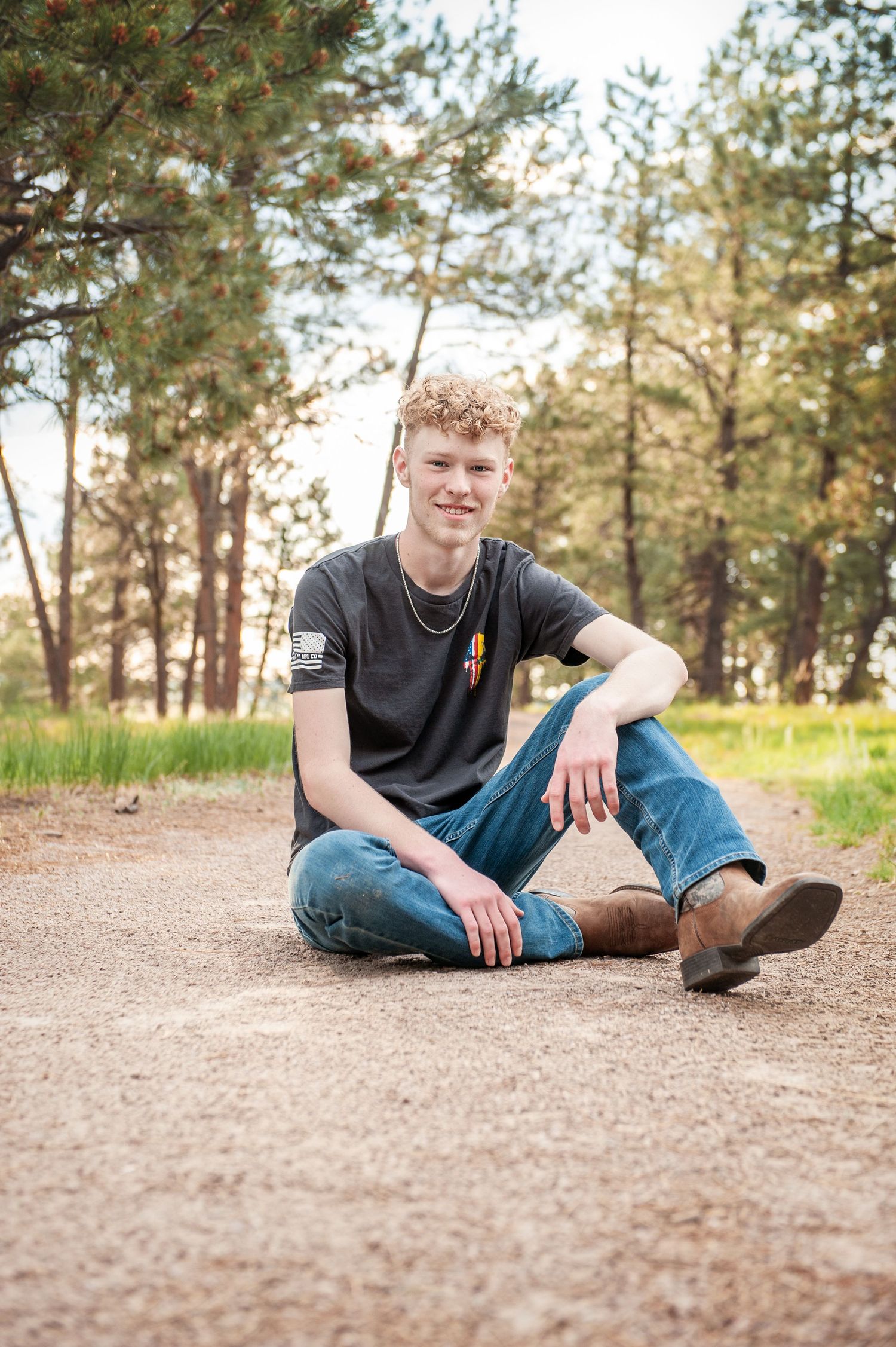 Person sitting cross-legged on dirt path with pine trees in background.