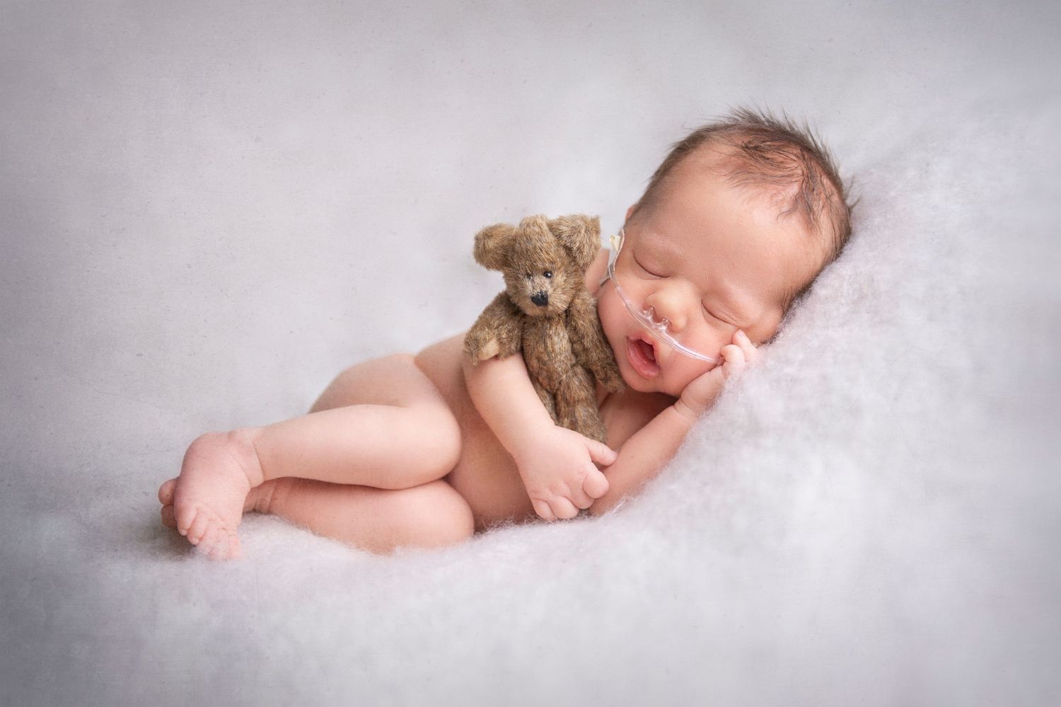 Sleeping newborn cuddling small teddy bear on soft white blanket.