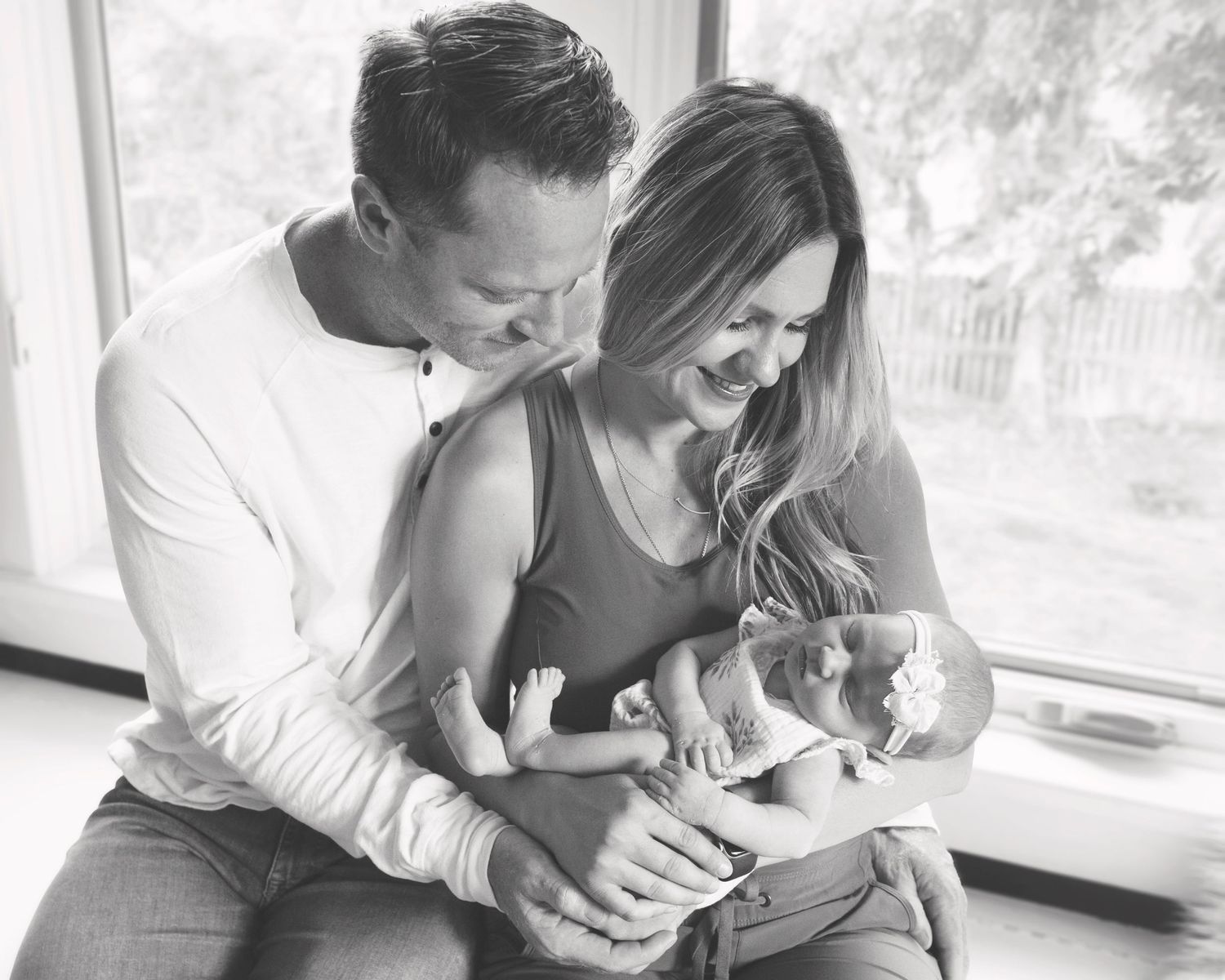 Intimate black and white portrait of parents tenderly holding and gazing at their newborn baby by a bright window.