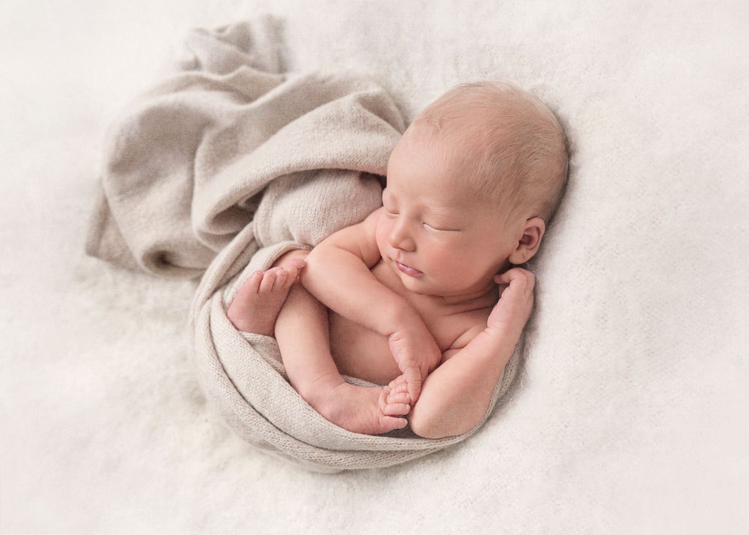 A peaceful sleeping newborn baby curled up on soft beige bedding.
