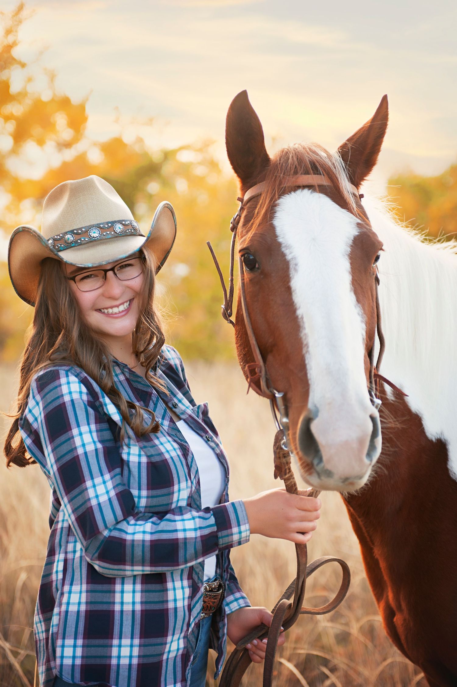 Western-styled portrait with cowboy hat and plaid shirt next to paint horse in autumn setting.