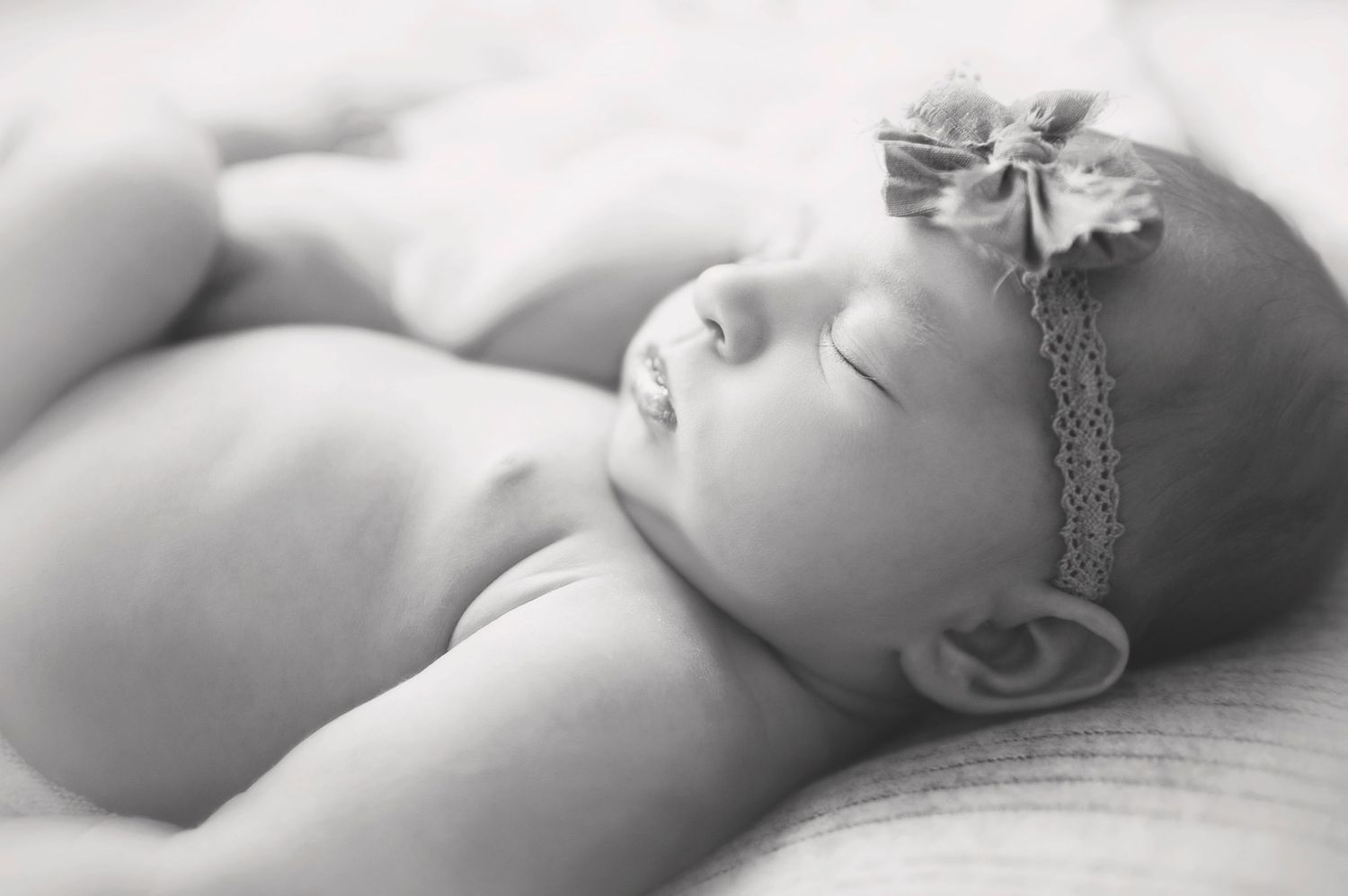 Black and white portrait of sleeping newborn with lace headband on soft textured bedding.