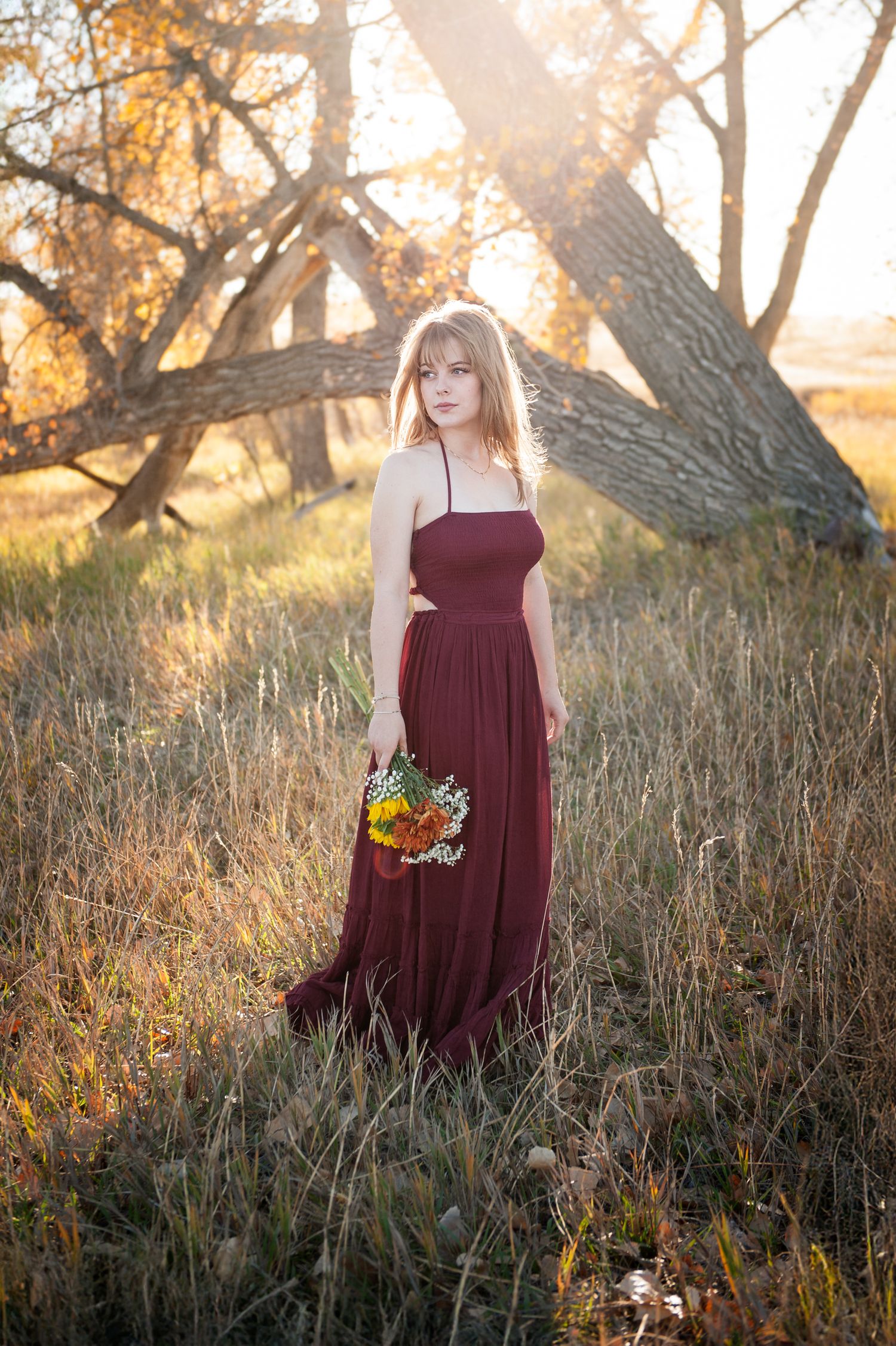 Person in burgundy dress holding flowers in sunlit autumn field.