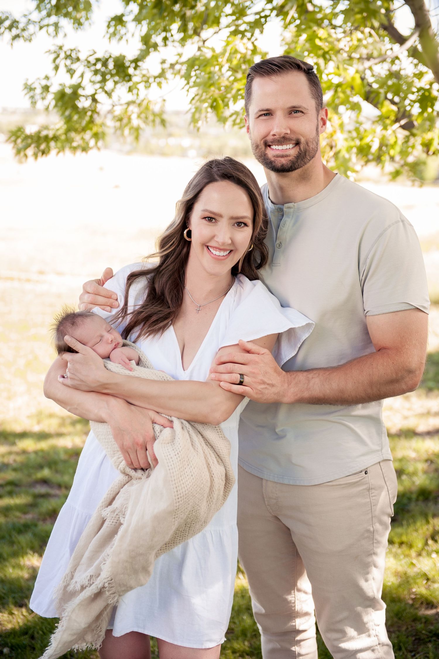 A sunny outdoor family portrait with a newborn wrapped in a cream blanket.