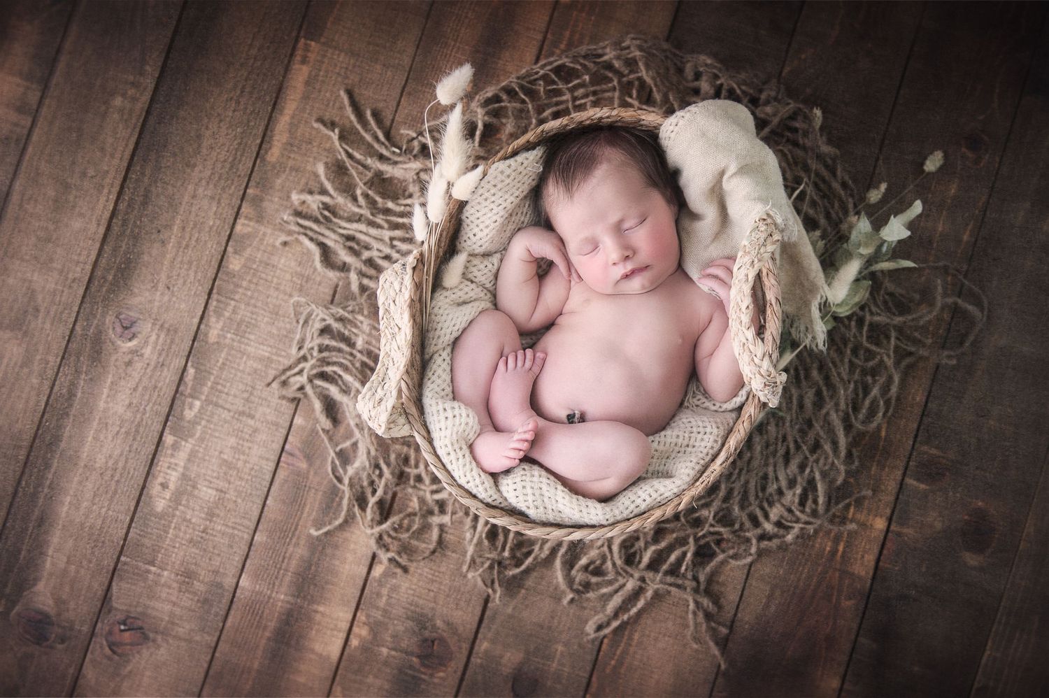 Overhead view of sleeping newborn in rustic basket with twigs and white fabric on wooden floor.