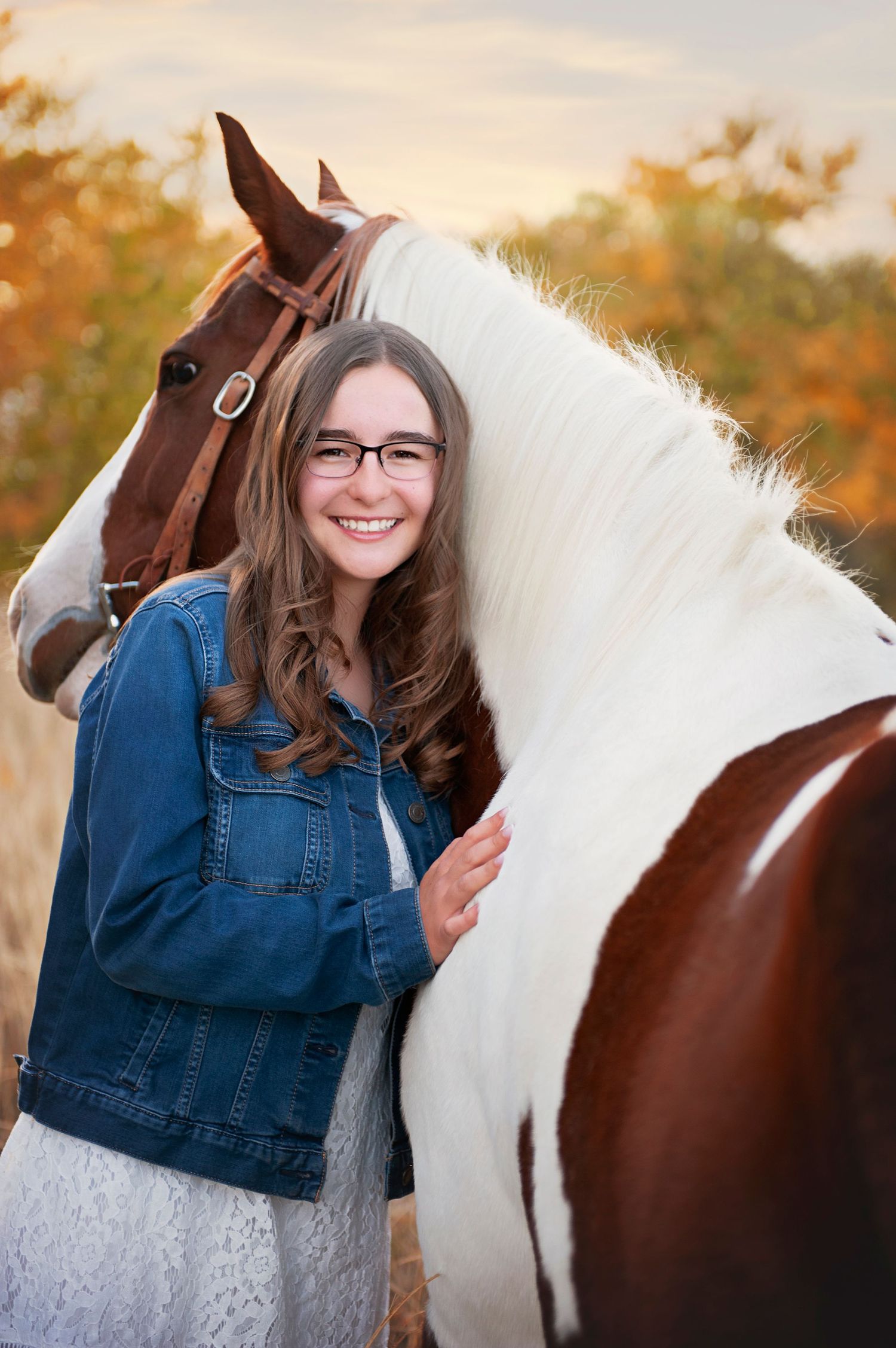 Person in denim jacket stands next to brown and white horse against autumn foliage.