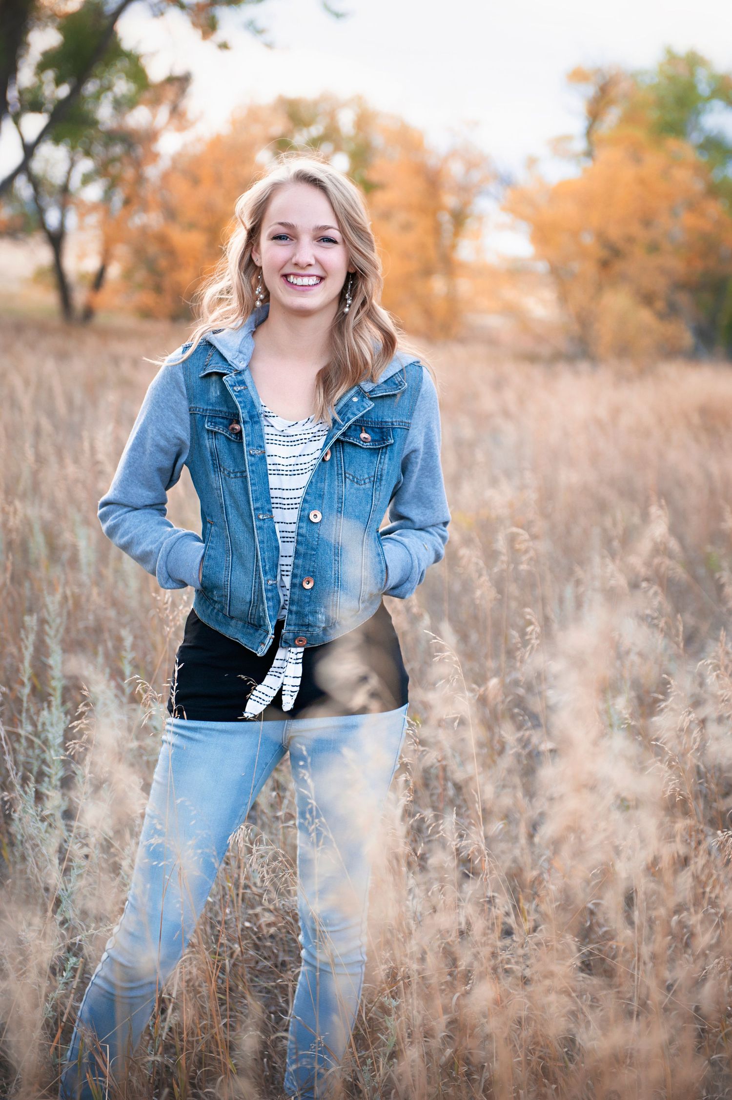 Person wearing denim jacket and jeans poses confidently in autumn field with dried grass.