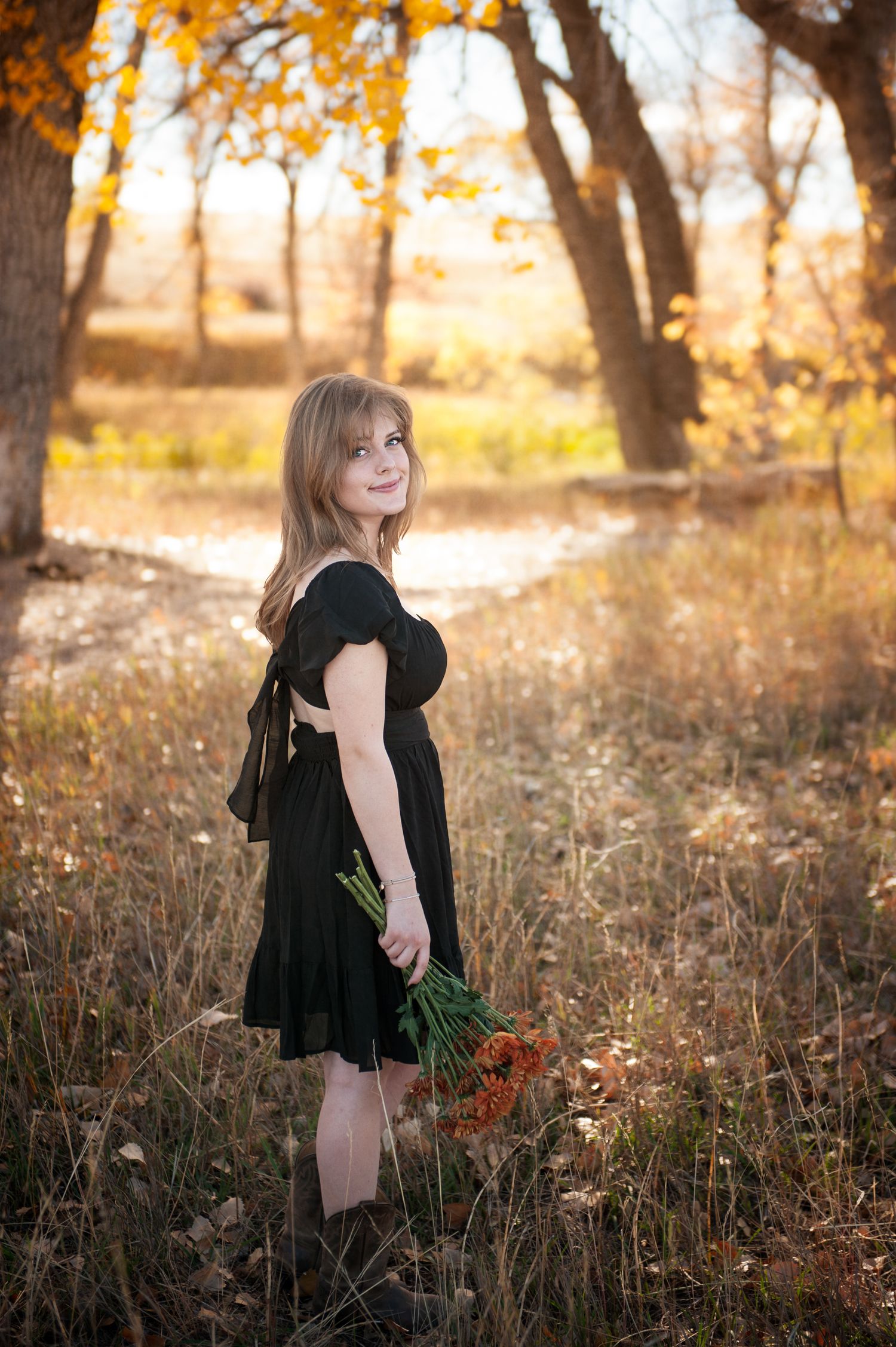 Full length portrait in black dress holding flowers in autumn woodland setting.