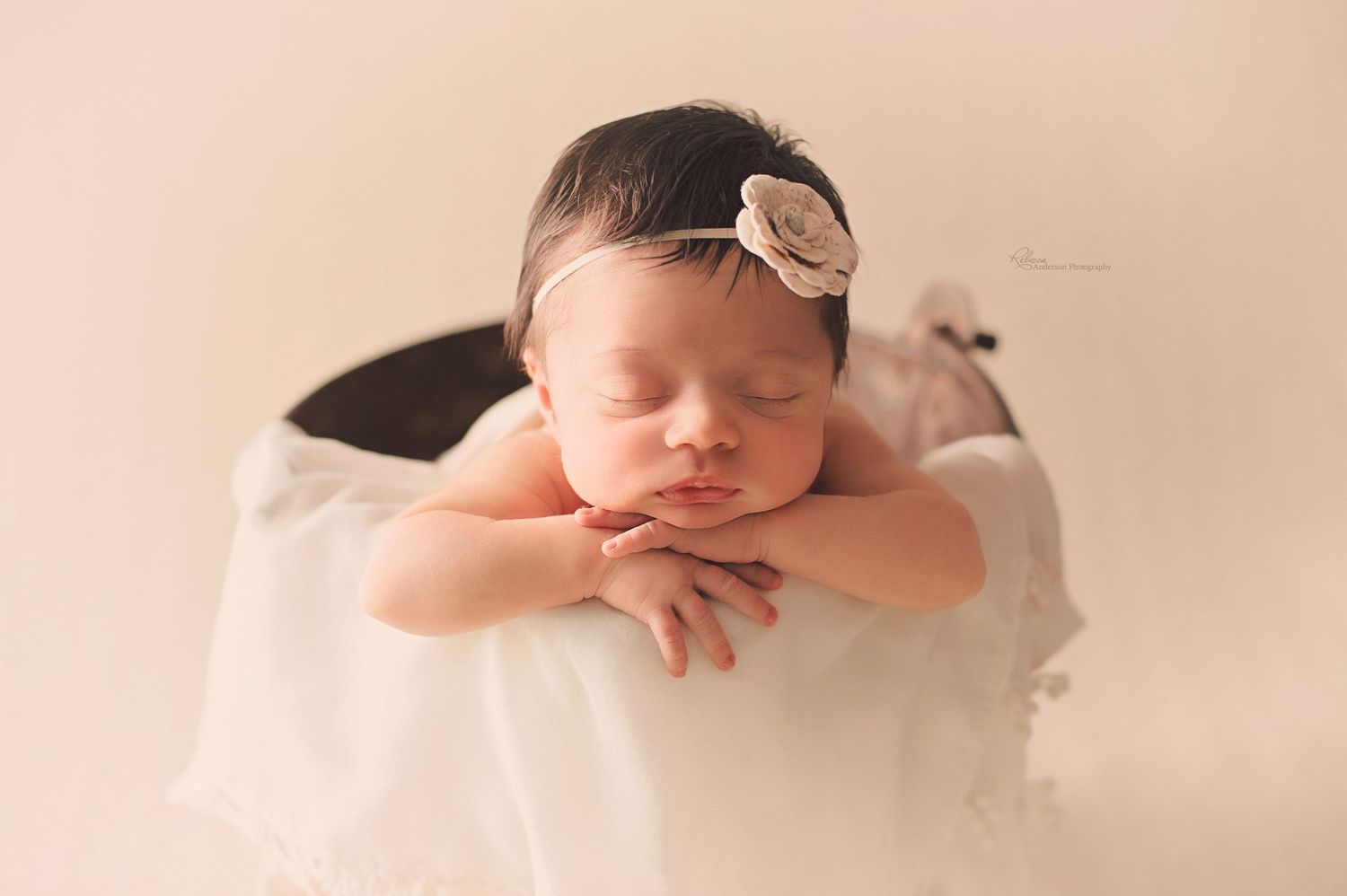 Sleeping baby resting on folded arms atop white fabric with delicate hair accessory.