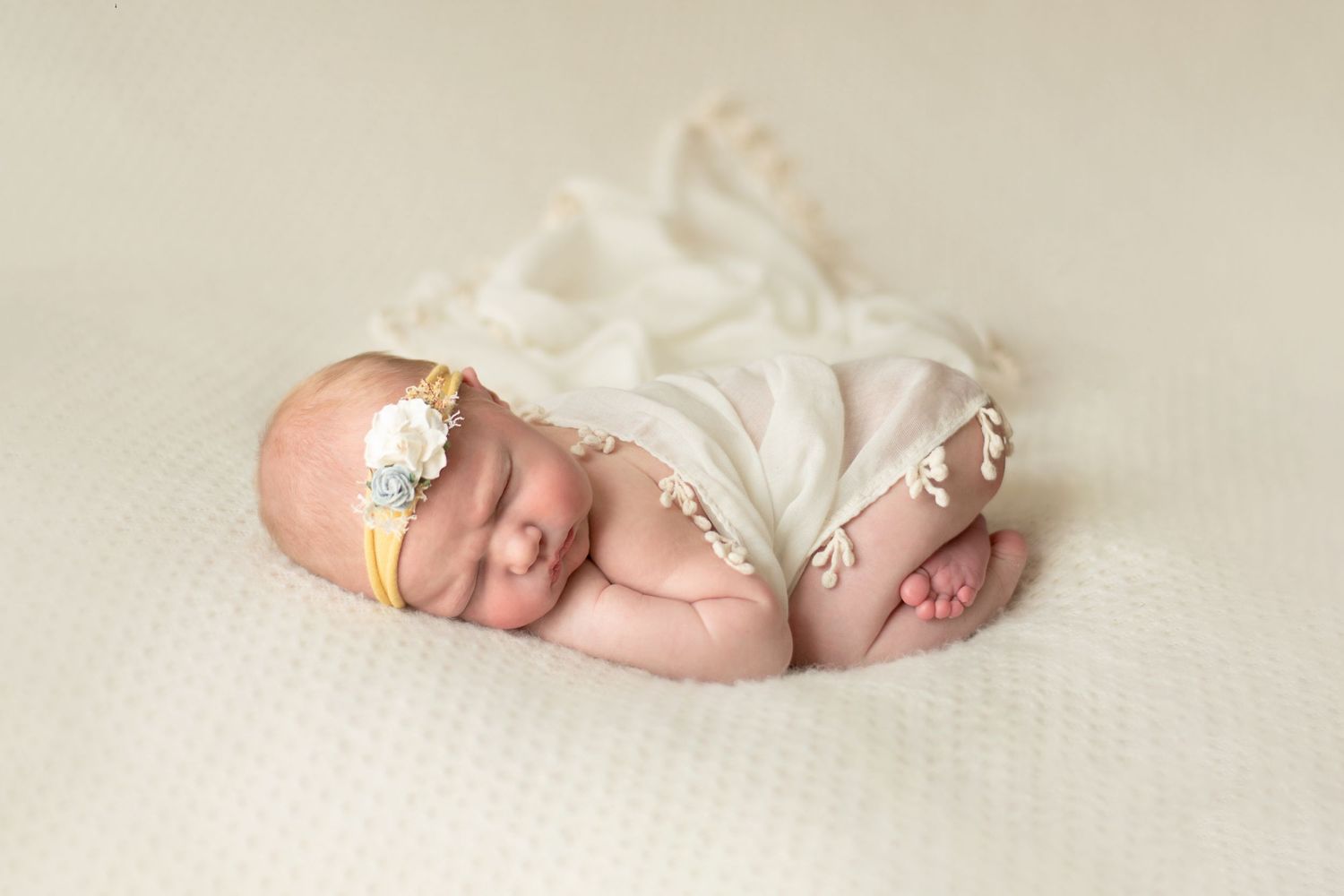 Sleeping newborn in soft white fabric wearing delicate floral headband lying on textured cream backdrop.