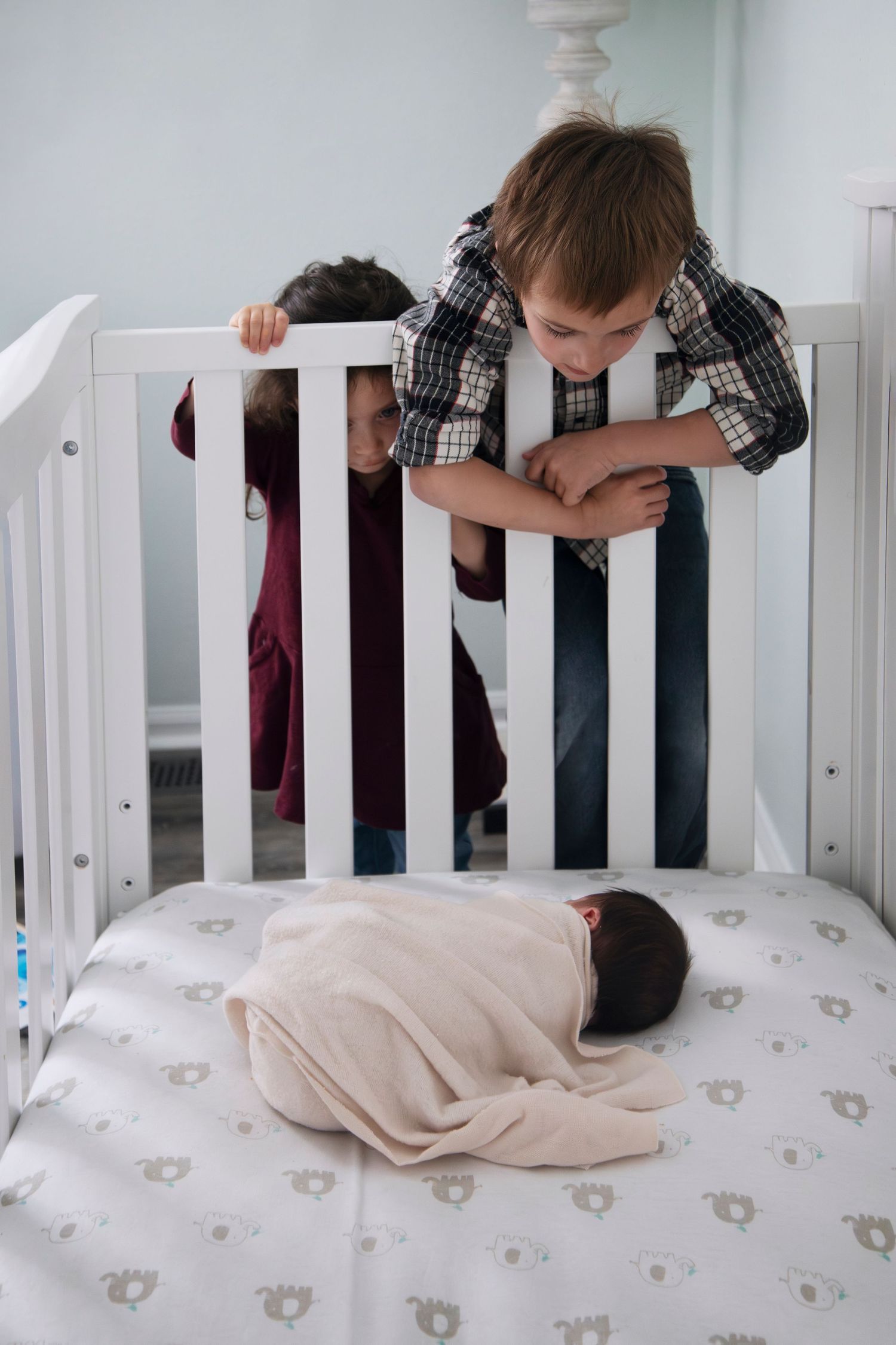 Young child peering through white crib railings at sleeping baby on patterned mattress.