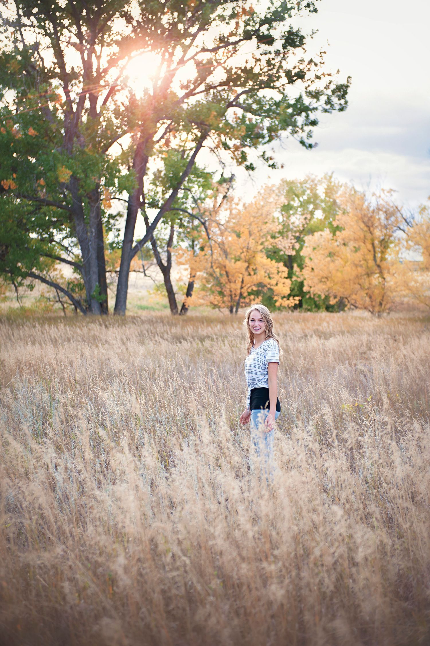 Full-length portrait standing in tall golden grass with sunlit trees.