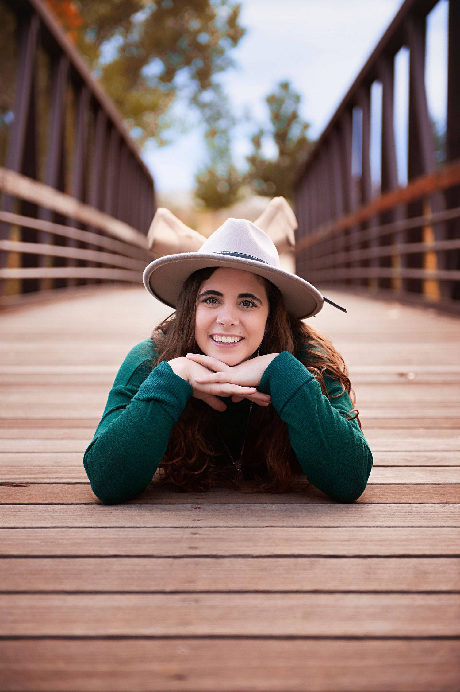 Person in a green sweater and beige hat lying on a wooden bridge with autumn scenery.