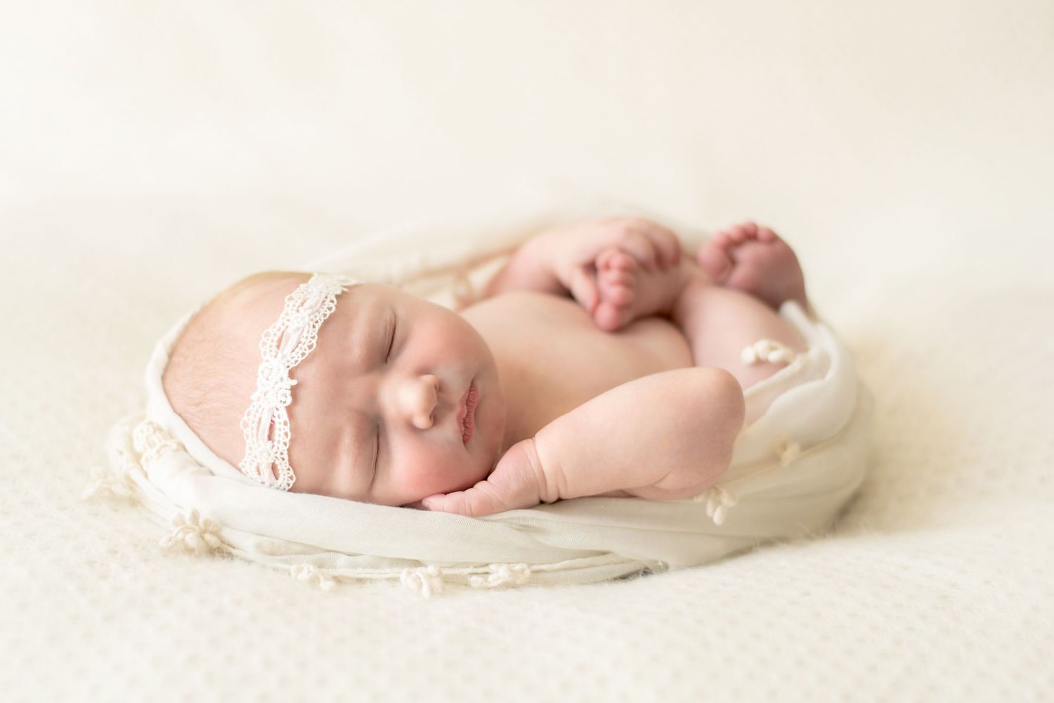 Peaceful newborn with lace headband sleeping on cream colored fabric.