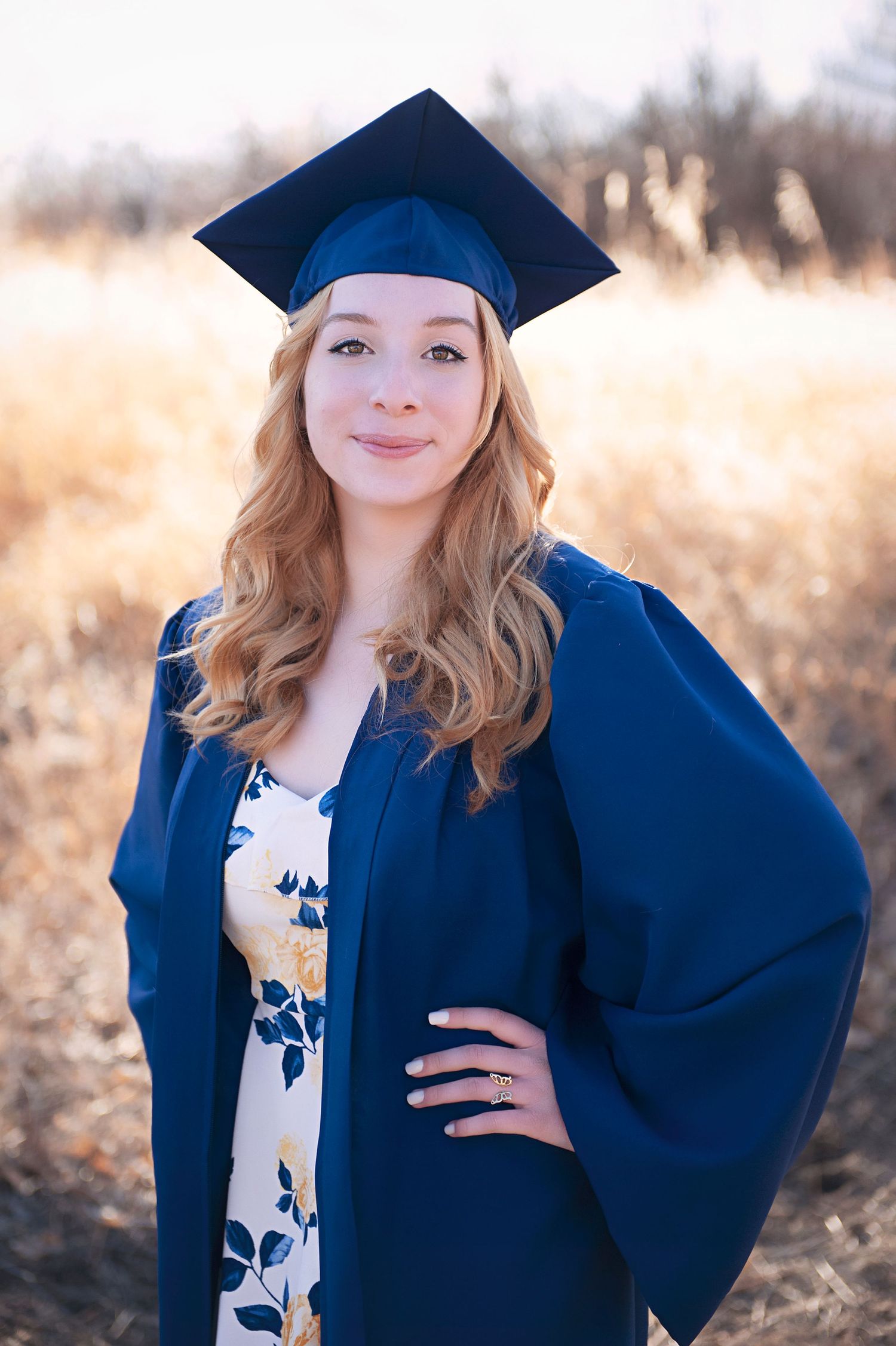 Graduate in blue cap and gown poses outdoors against blurred golden field background.