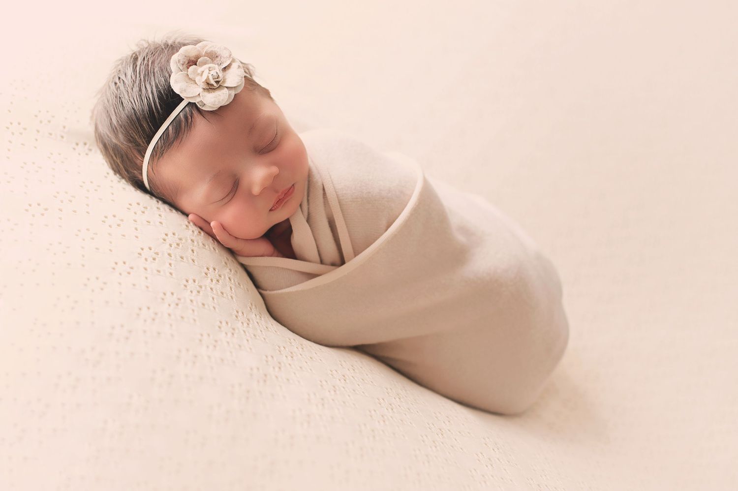Sleeping newborn wrapped in beige swaddle with delicate floral headband on cream background.