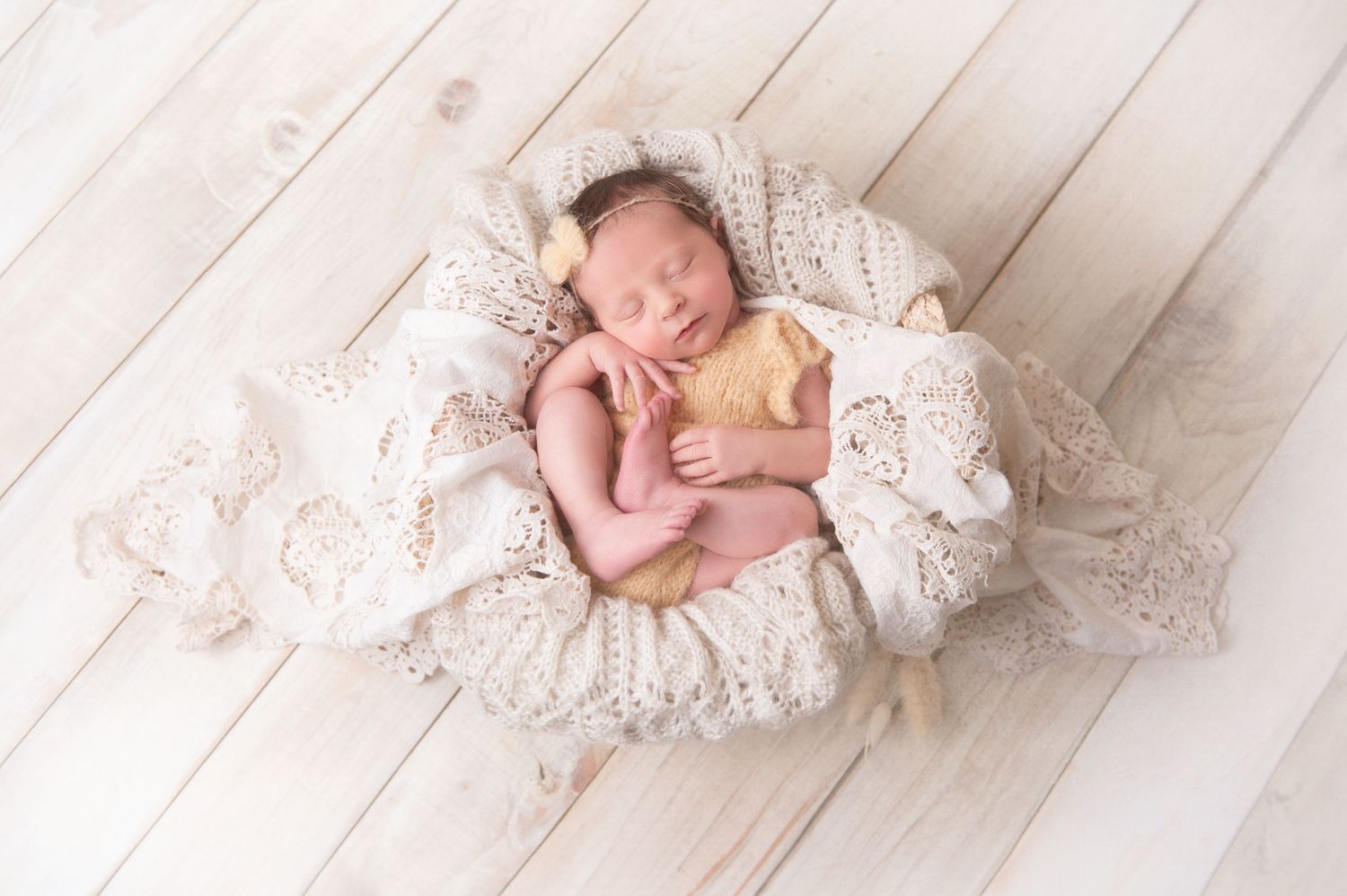 Sleeping newborn nestled in delicate lace fabric on white wooden backdrop.