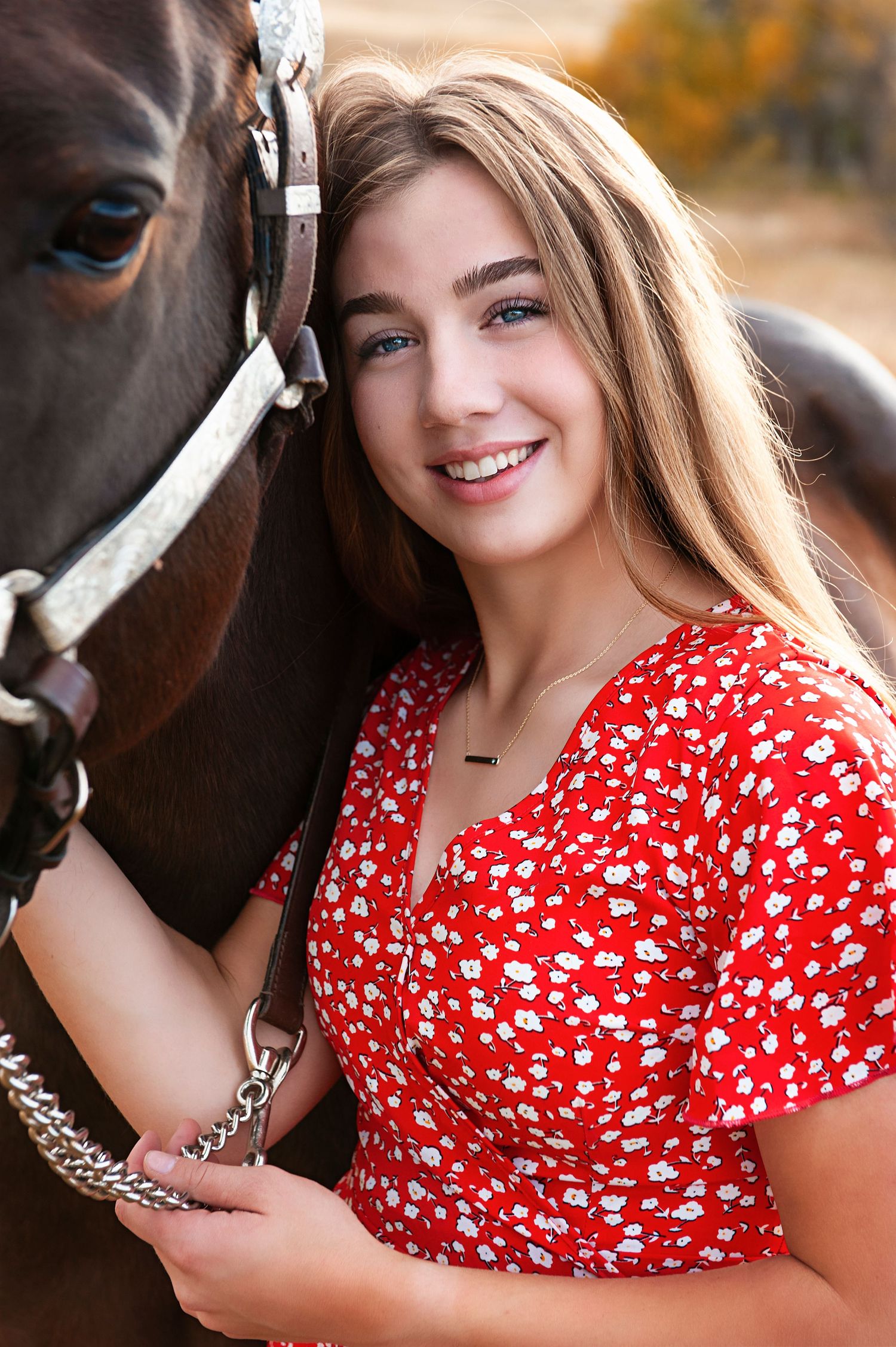Woman in red polka dot dress standing next to a brown horse with a bridle in an outdoor fall setting.