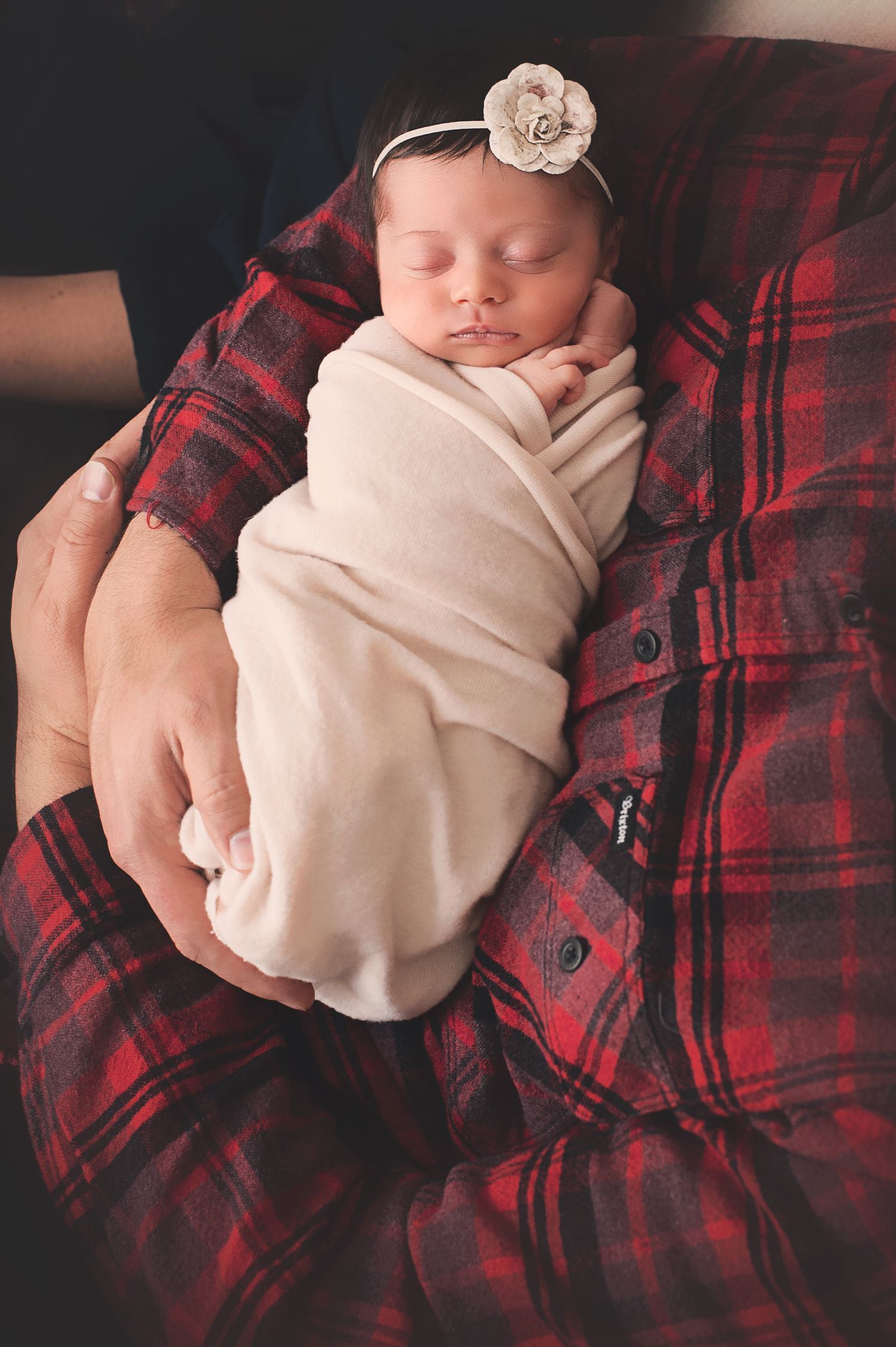 Sleeping newborn wrapped in white with flower headband resting on red plaid flannel.