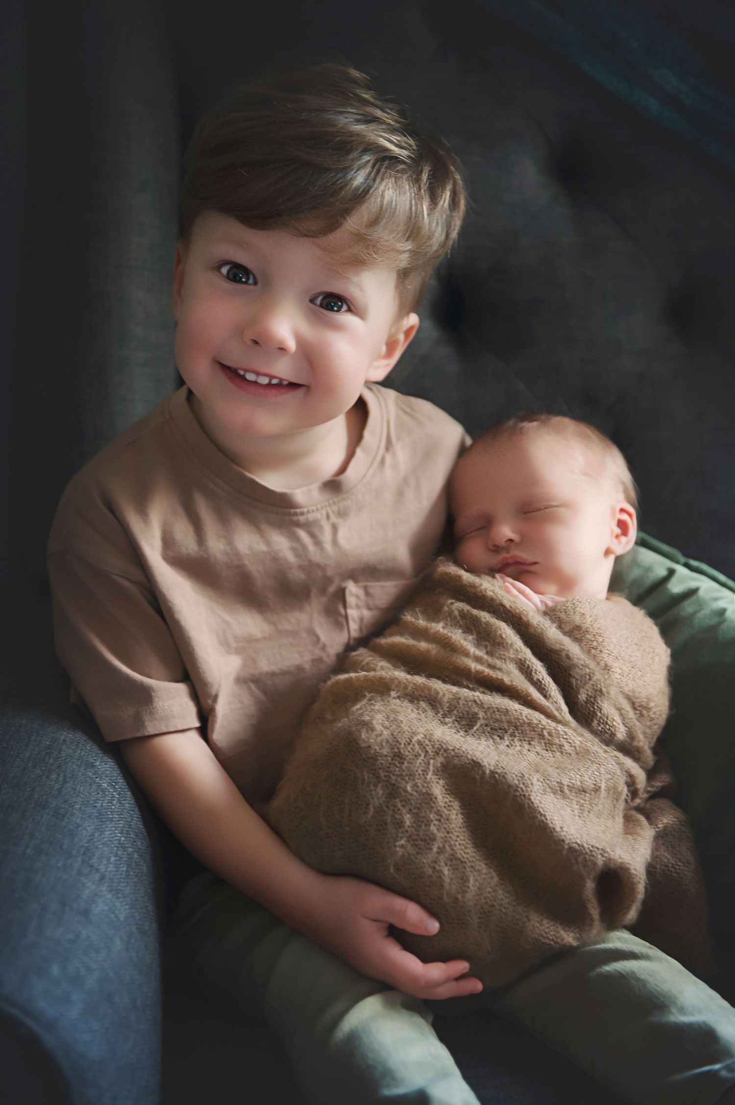 A toddler in beige holds a swaddled newborn baby while sitting together in a dark studio setting.