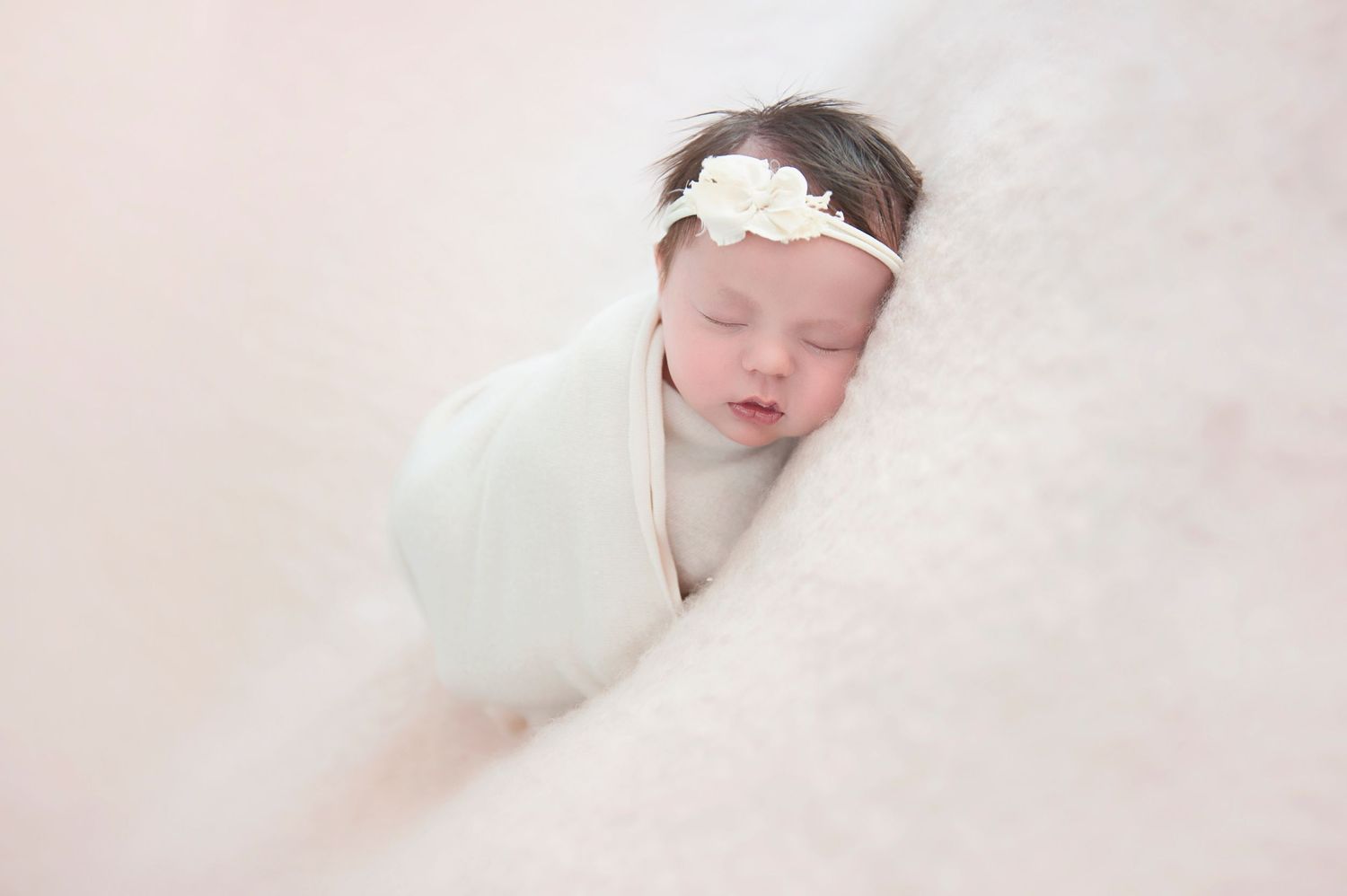Peaceful newborn swaddled in white wrap with floral headband on light fabric background.
