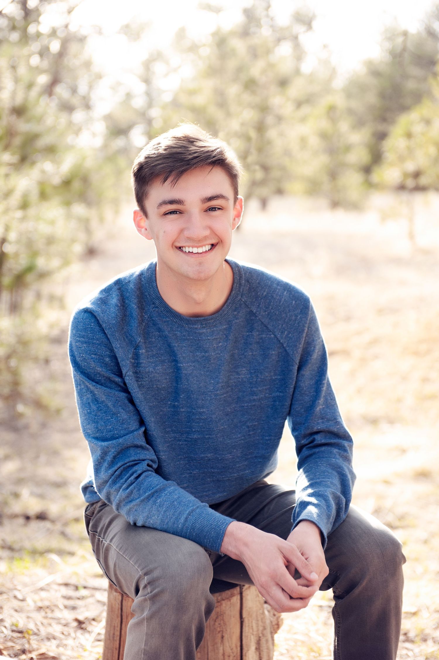 Person sitting casually outdoors in blue sweater with soft natural lighting and pine trees in background.