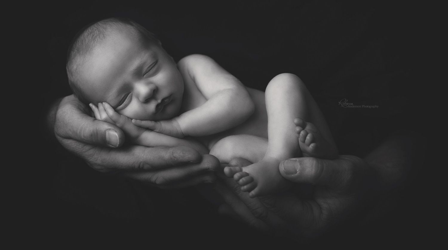Black and white image of a sleeping newborn cradled in gentle hands.