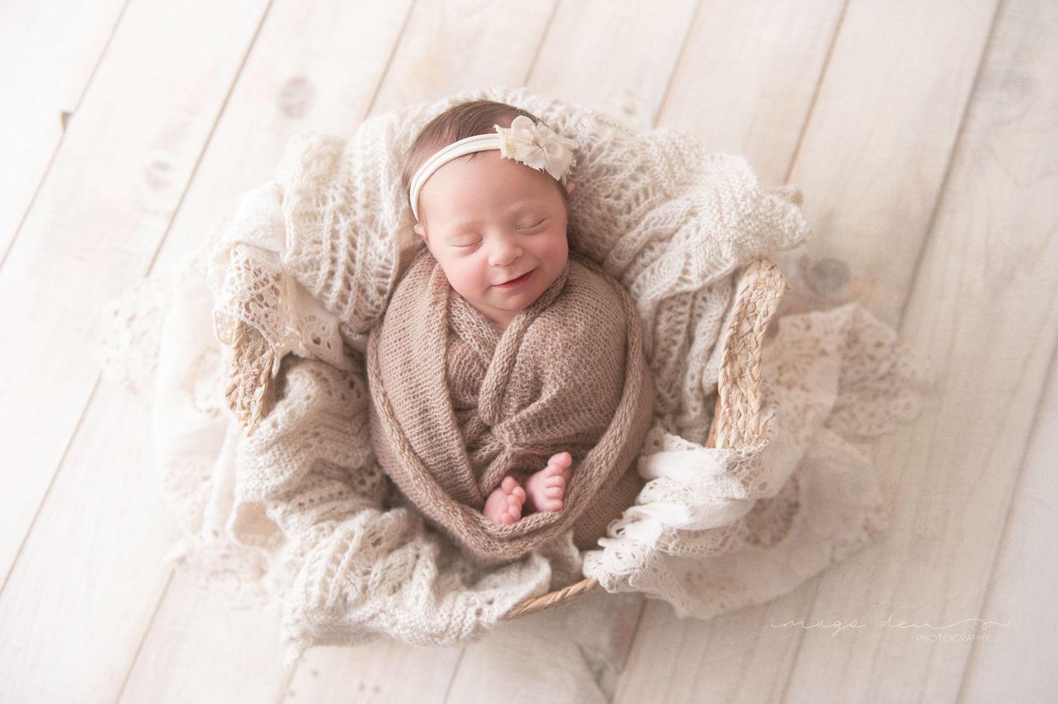 Newborn baby sleeping peacefully in tan wrap surrounded by lacy fabric on wooden floor.